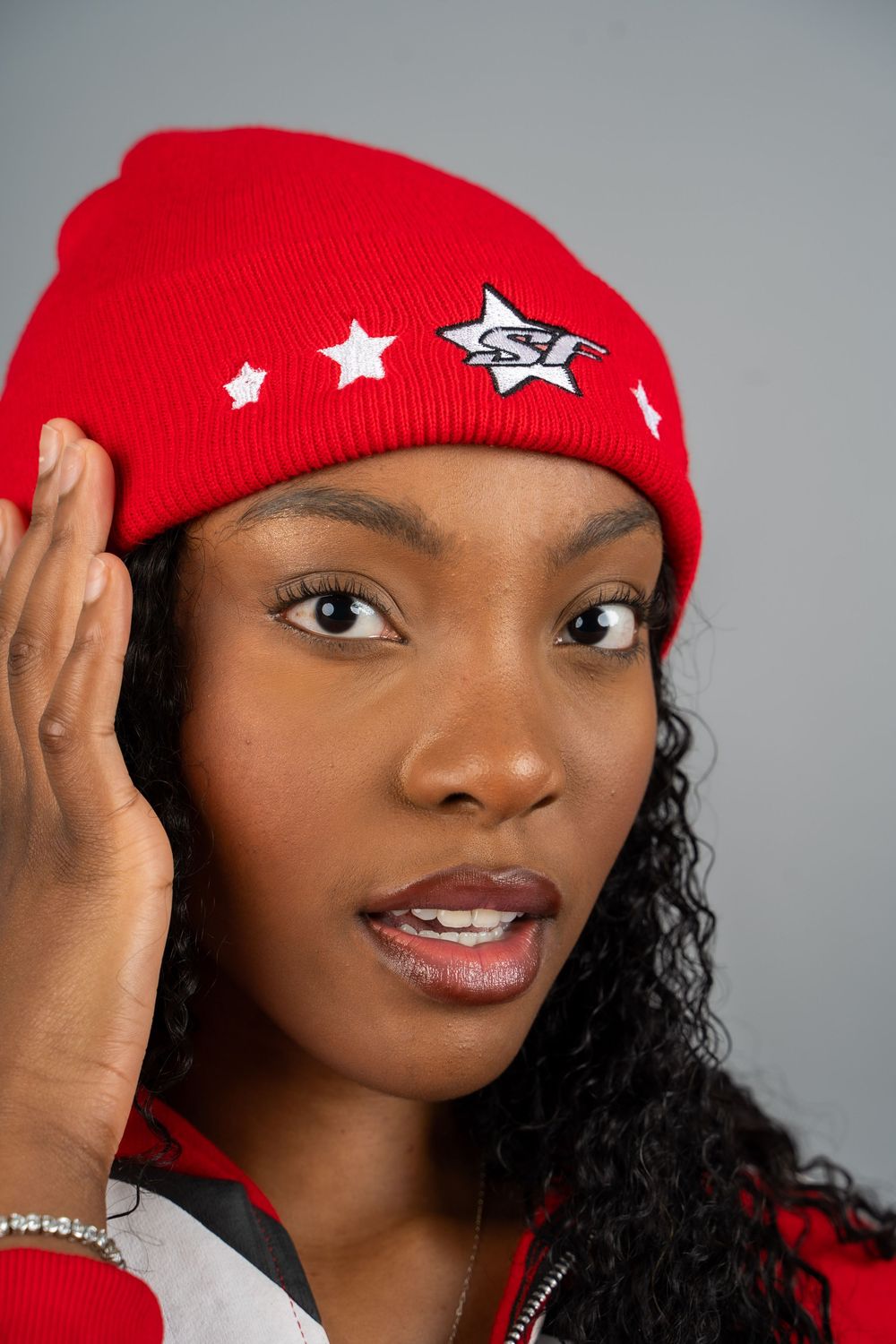 Fashion model wearing a red beanie with star decorations and natural curly hair against a gray studio background.