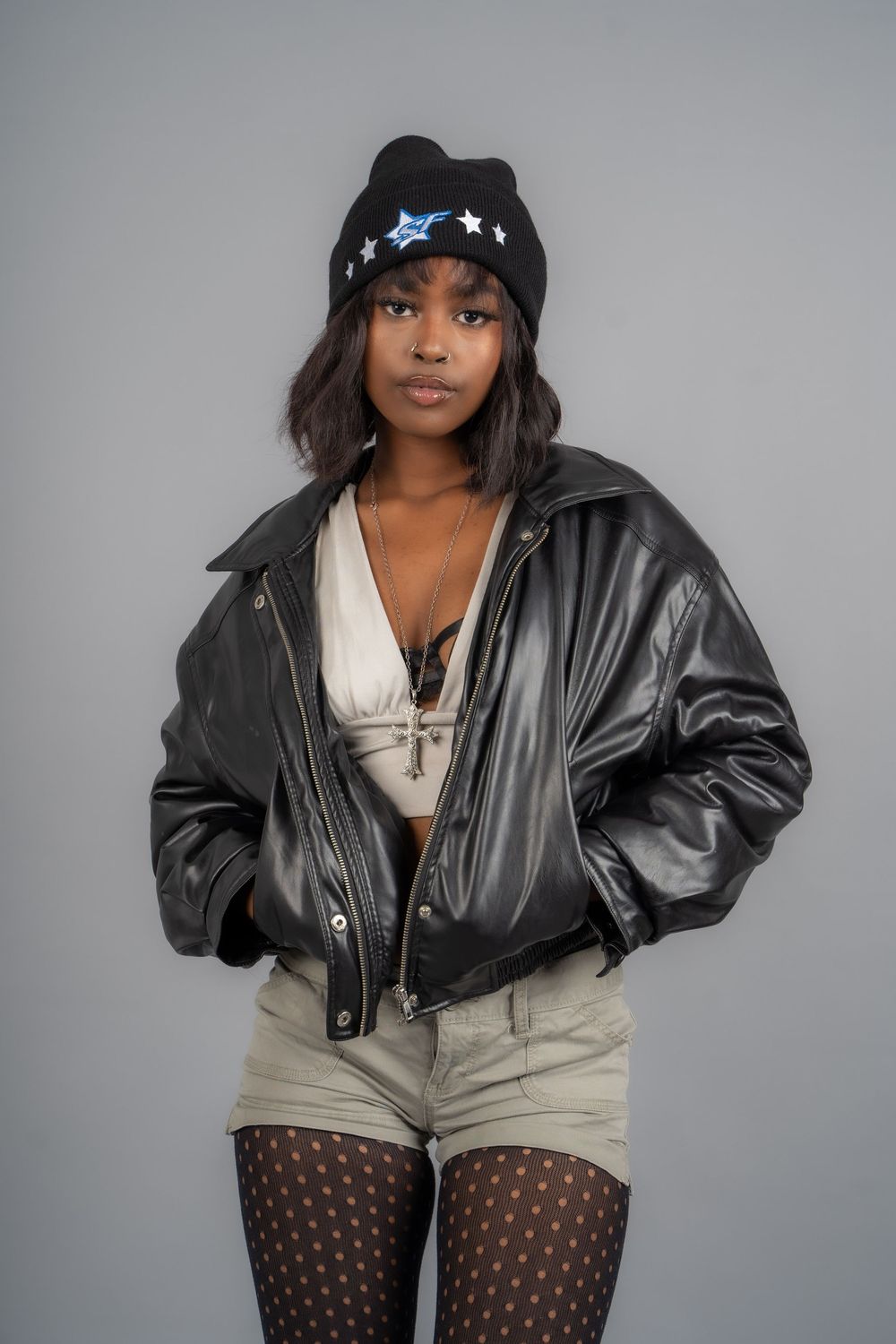 Urban fashion look with black leather jacket, beige shorts and polka dot tights in studio setting.