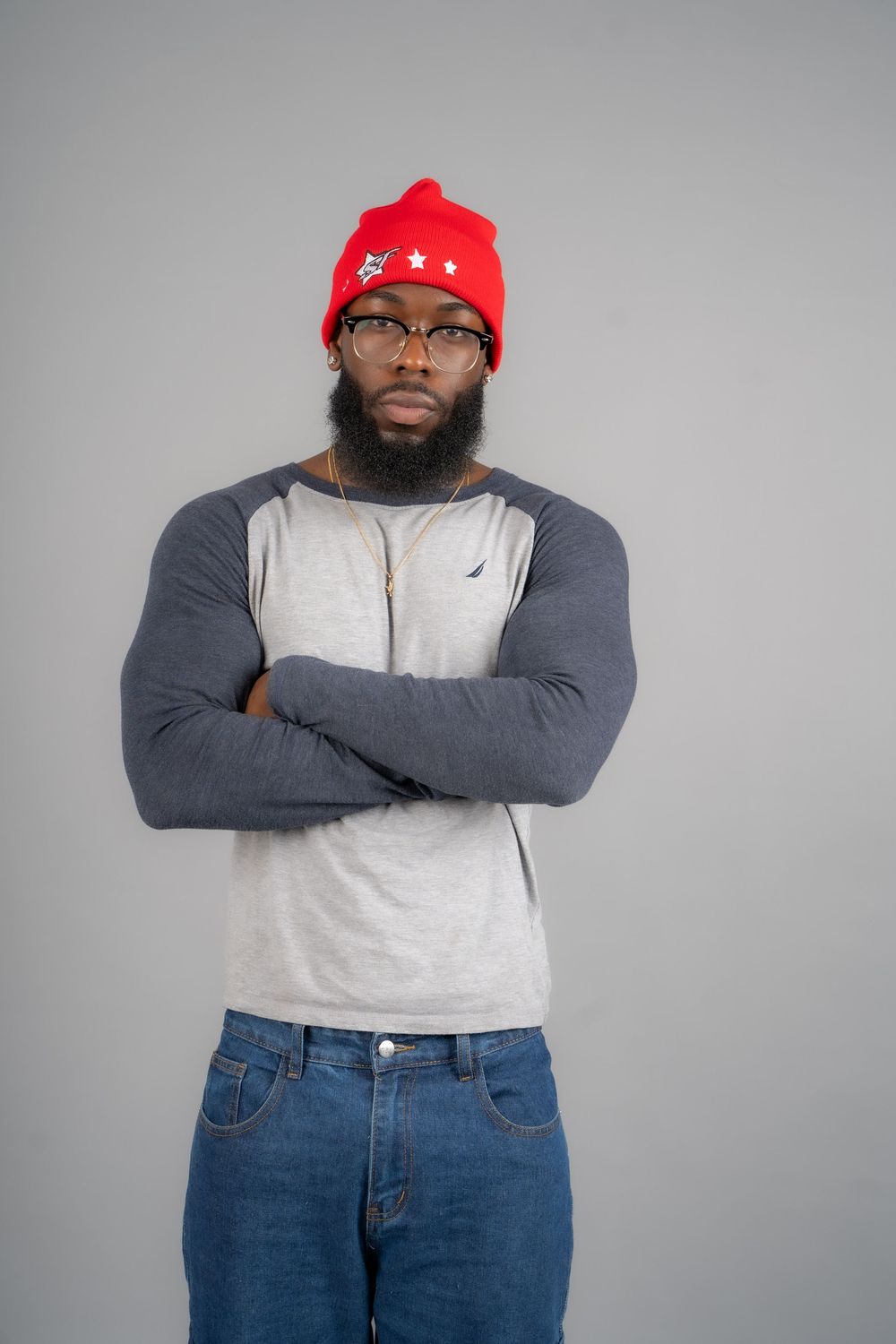 Person in red cap, gray sweater and jeans standing with crossed arms against gray background.