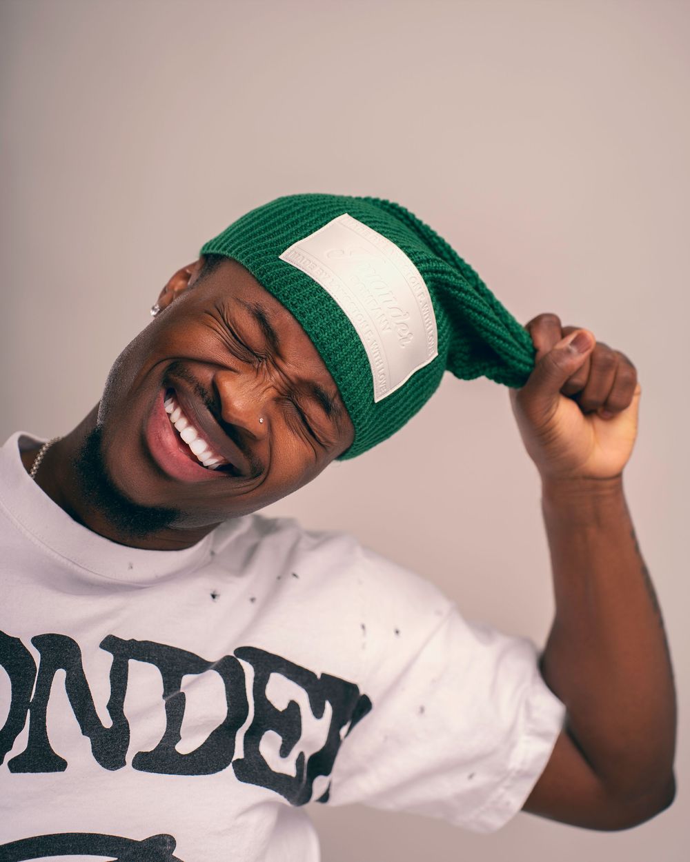 Person wearing a green and white beanie hat and white t-shirt smiling joyfully against neutral background.