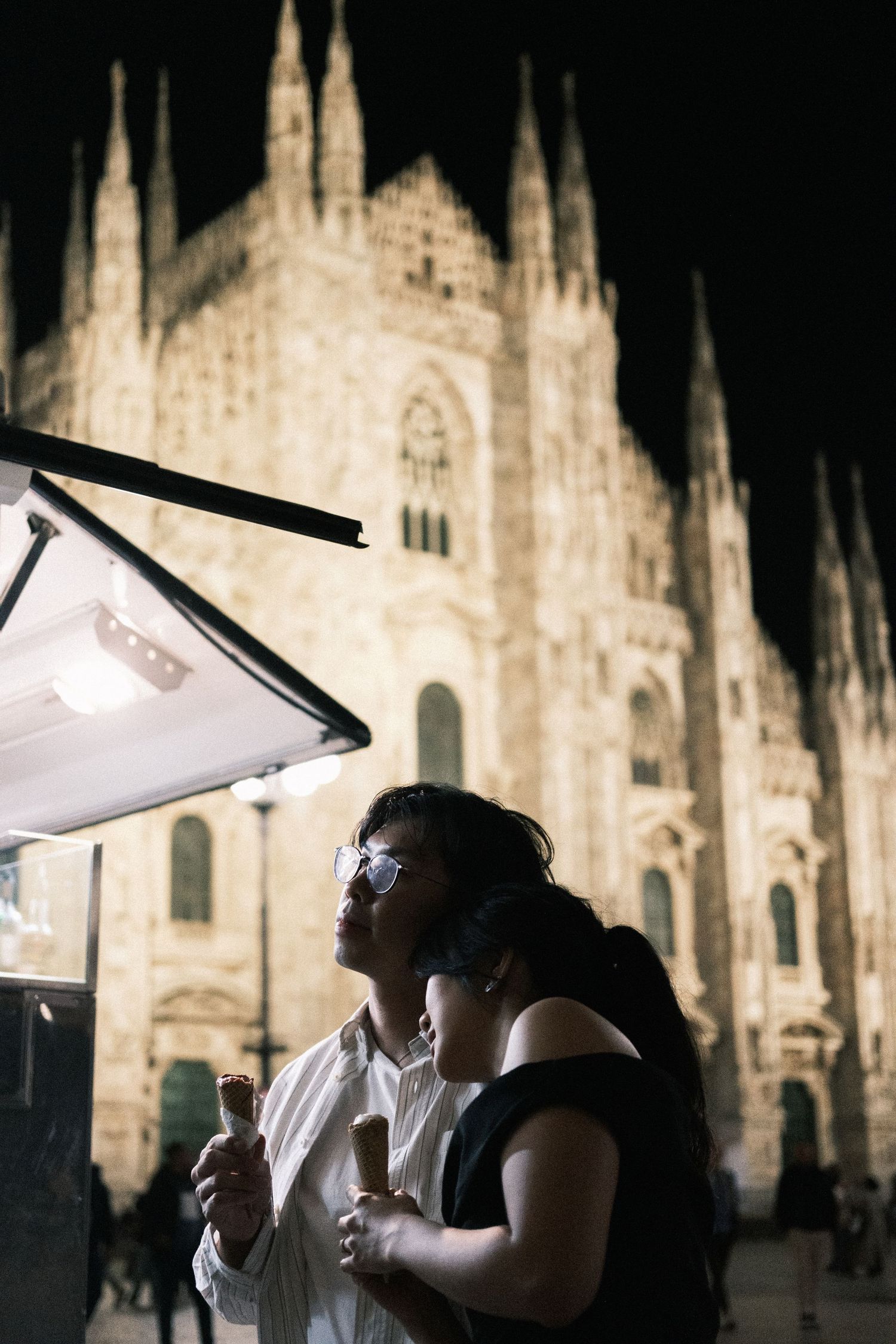 People enjoy gelato at night in front of the illuminated Milan Cathedral's Gothic facade in Italy.