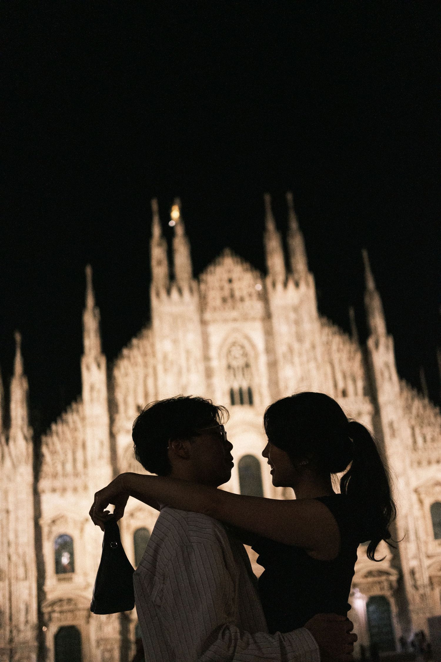 A romantic silhouette of a couple embracing in front of the illuminated Milan Cathedral at night.