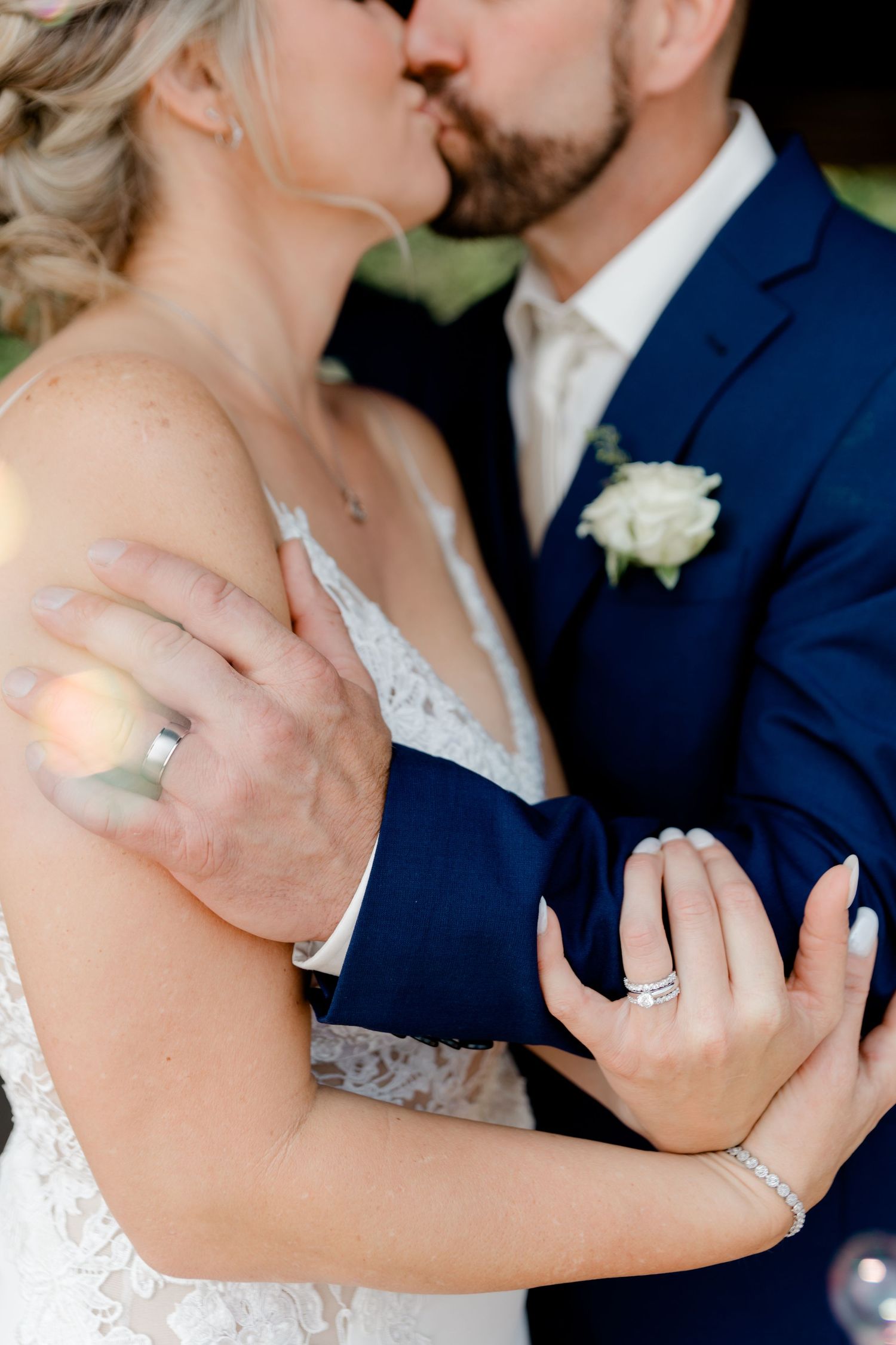 Bride and groom kissing while showcasing wedding rings, groom in navy suit with white rose boutonniere.