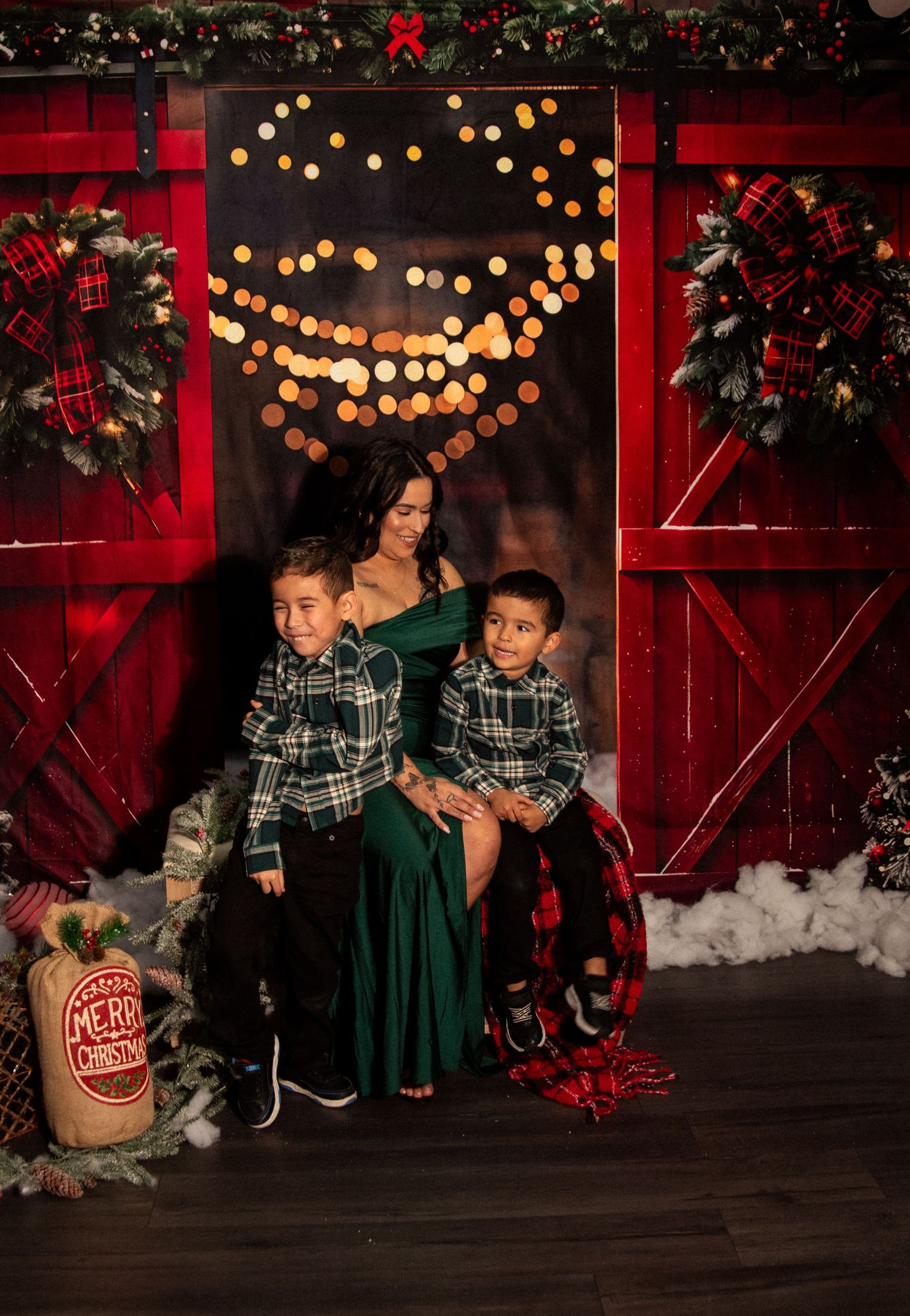 A festive holiday scene with green velvet dresses against a red barn backdrop with twinkling lights and Christmas decorations.