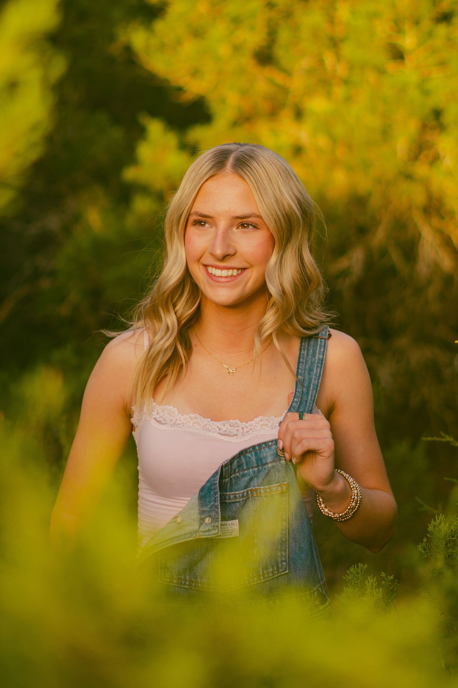 Person in overalls and pink top poses among yellow autumn foliage.