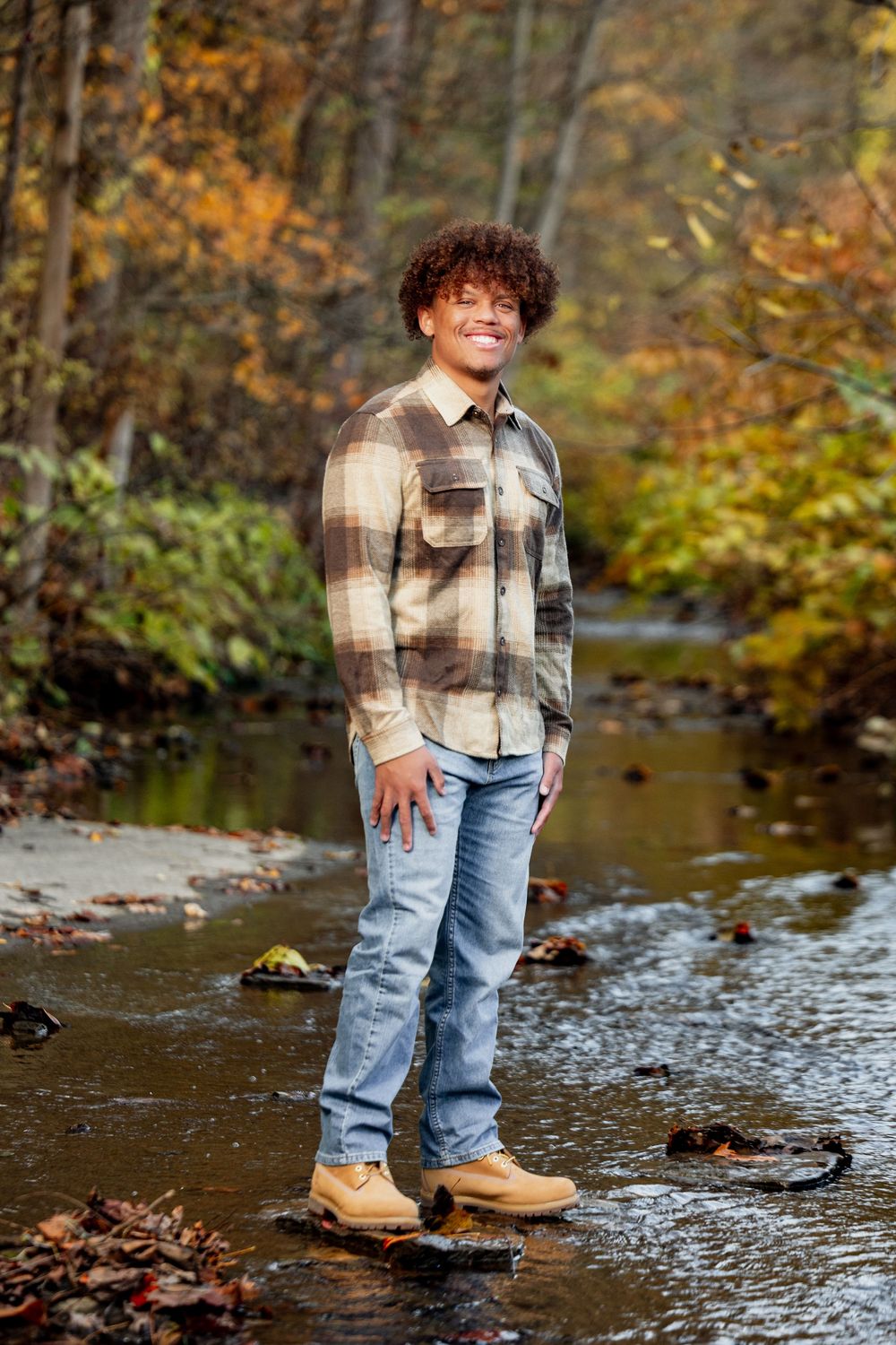 Child wearing plaid shirt and jeans stands in shallow creek during autumn with colorful fall foliage in background.