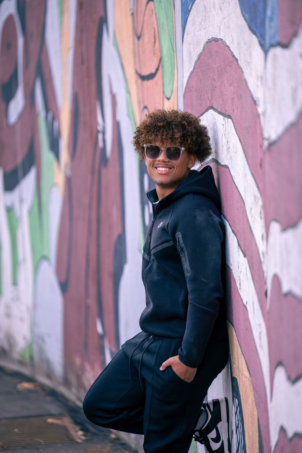 Person in black hoodie and sunglasses poses against colorful graffiti wall in urban setting.