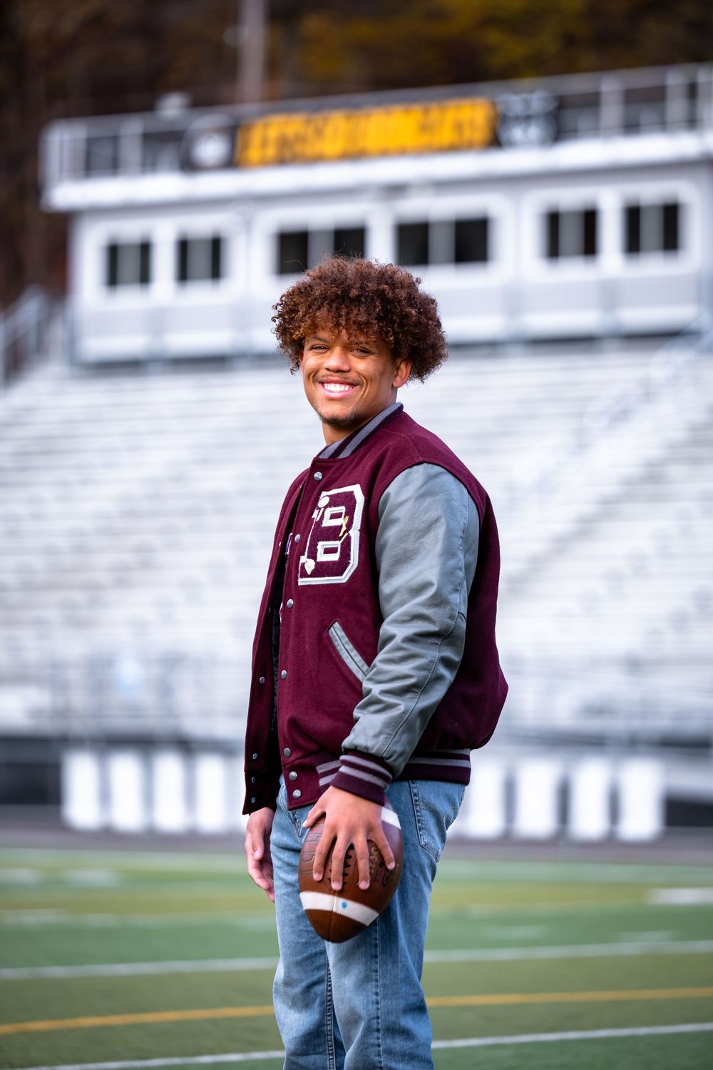 Young child wearing varsity jacket and jeans stands on football field with bleachers in background.