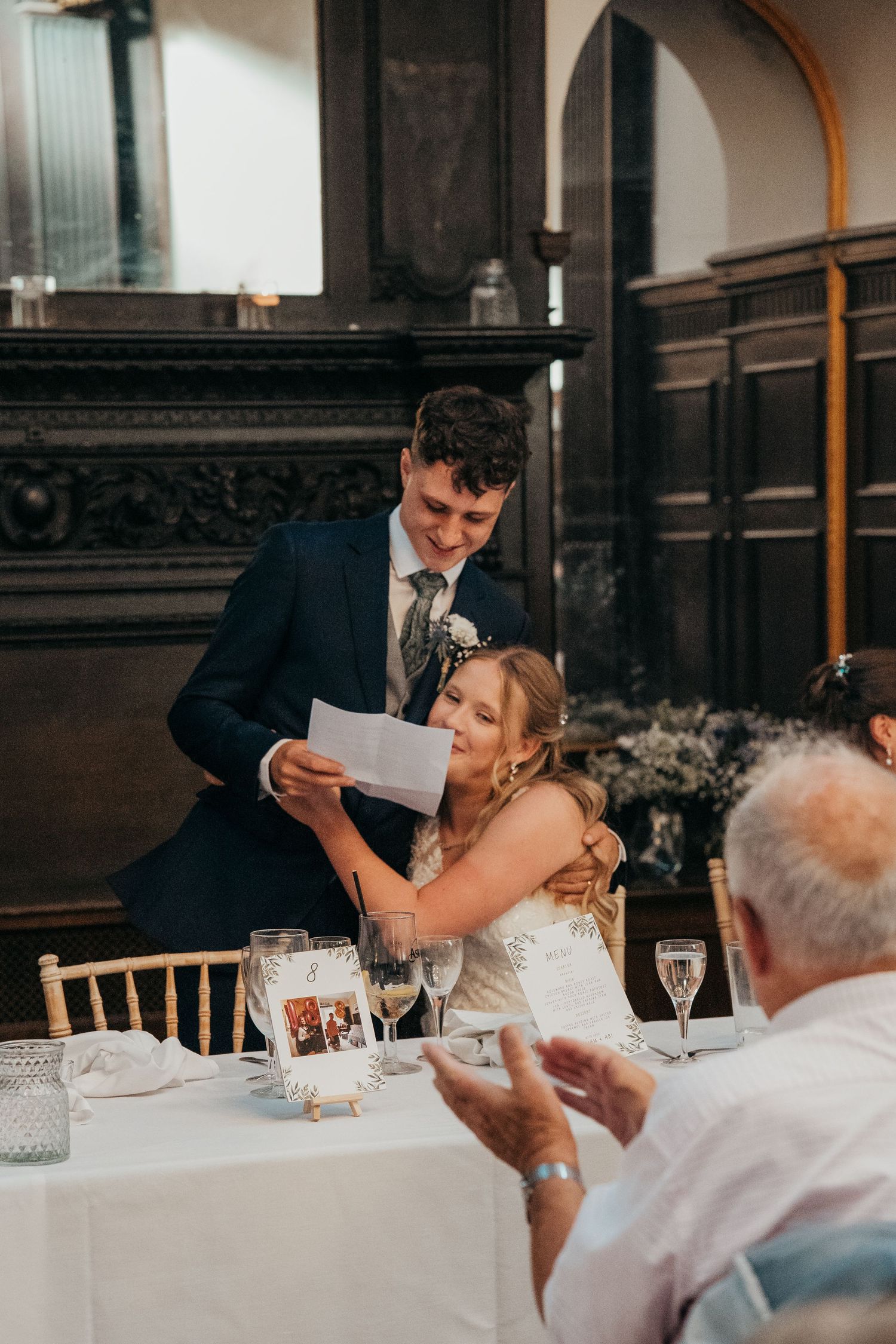 Wedding reception scene showing dining table with guests and speeches in elegant venue.
