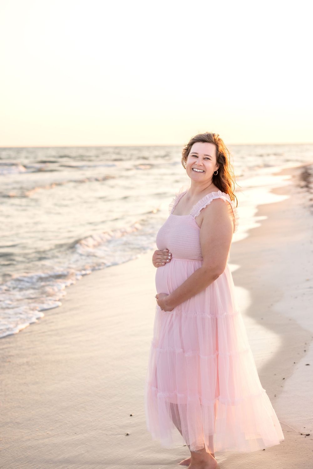 A person in a flowing pink dress walks along a sandy beach at sunset with waves gently rolling in.