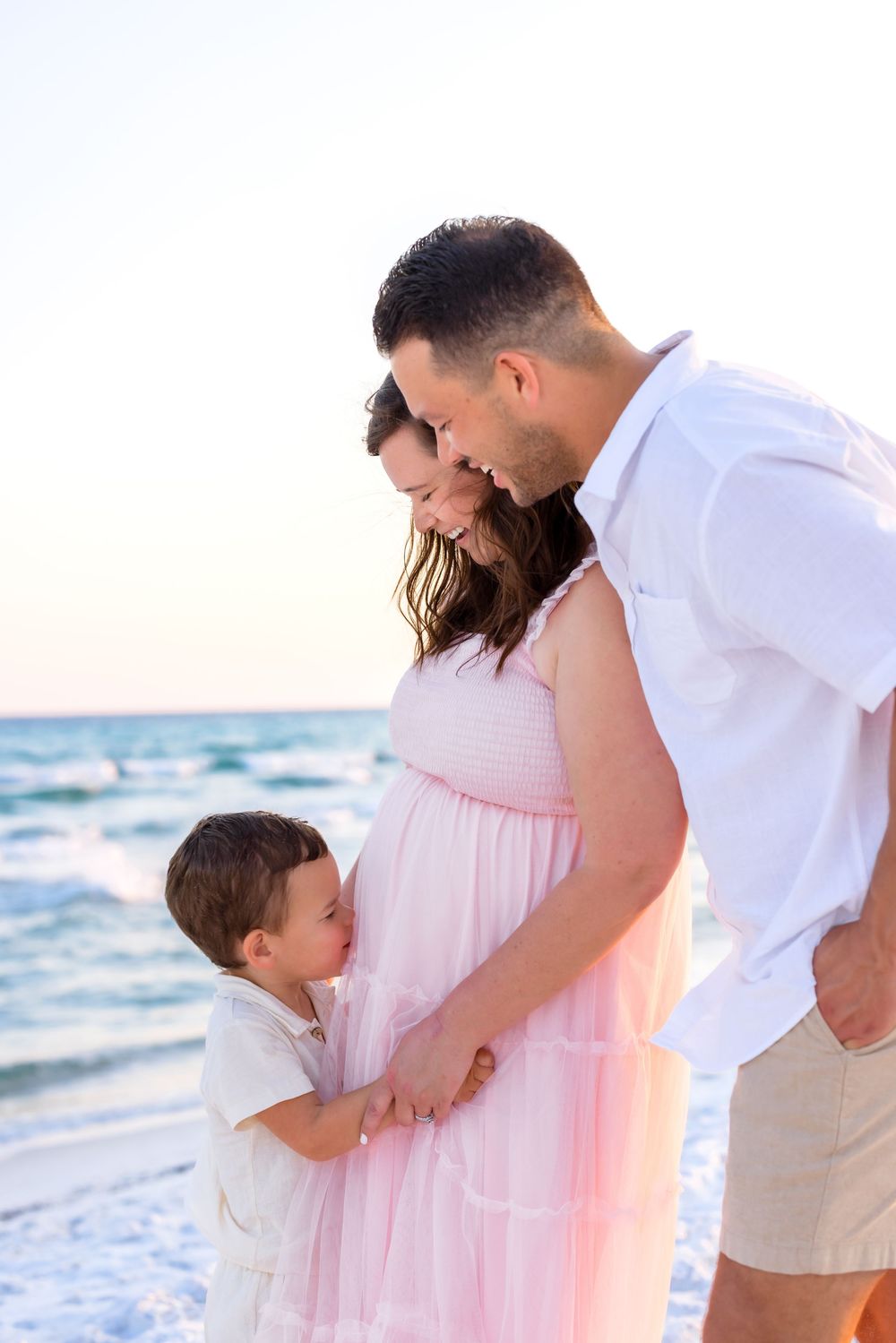 A family shares tender moments on a beach at sunset wearing white and pink outfits.