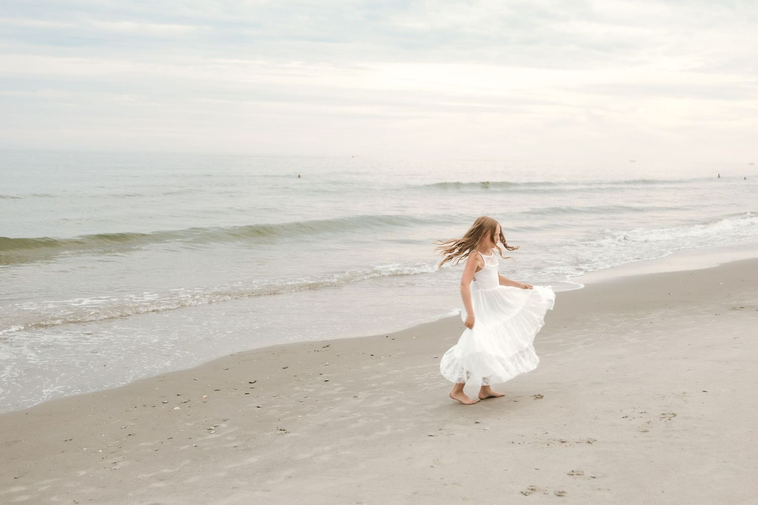 A bride in a flowing white dress walks along a serene beach shoreline at sunset.