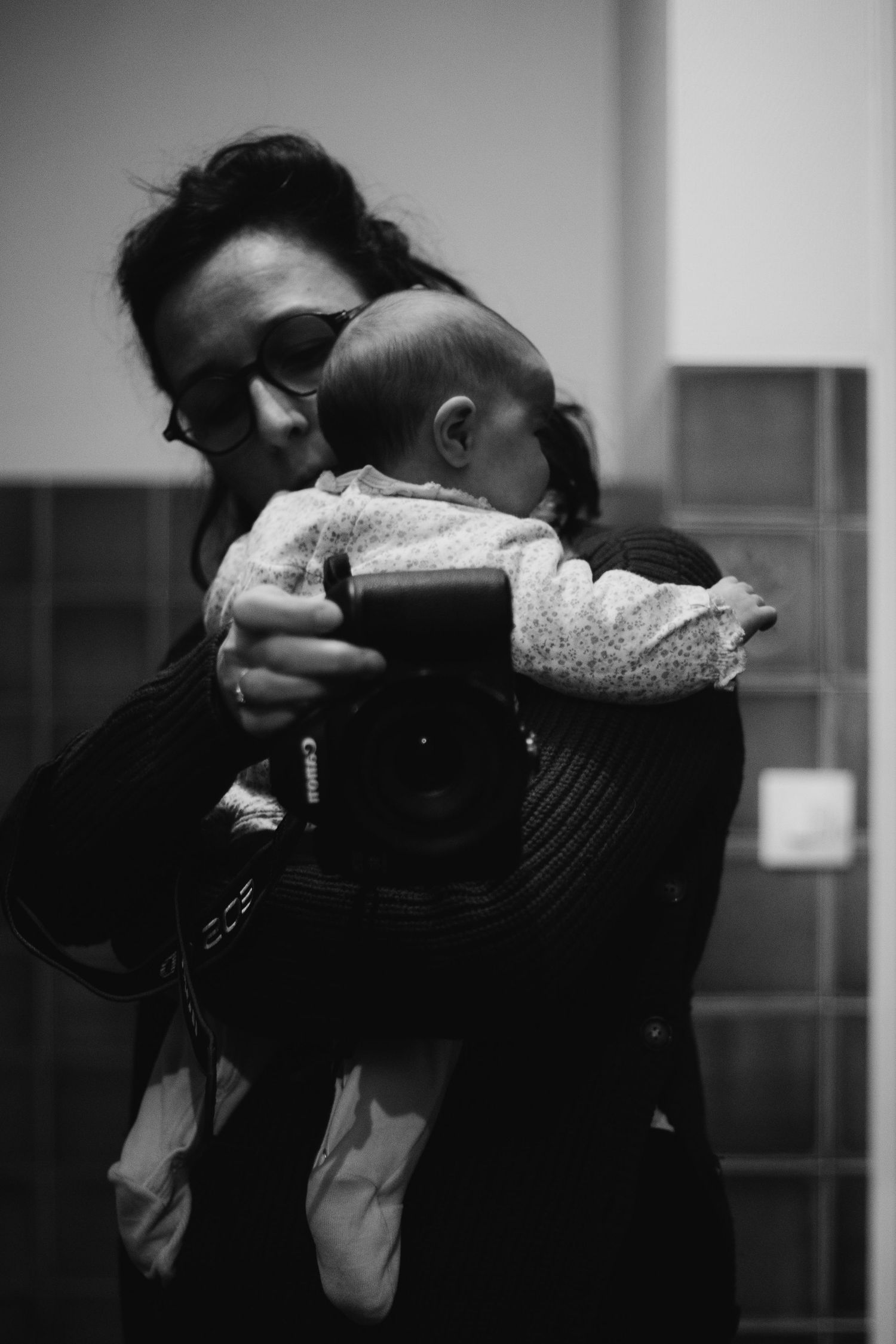 Black and white photo of woman holding newborn baby and Canon camera indoors.
