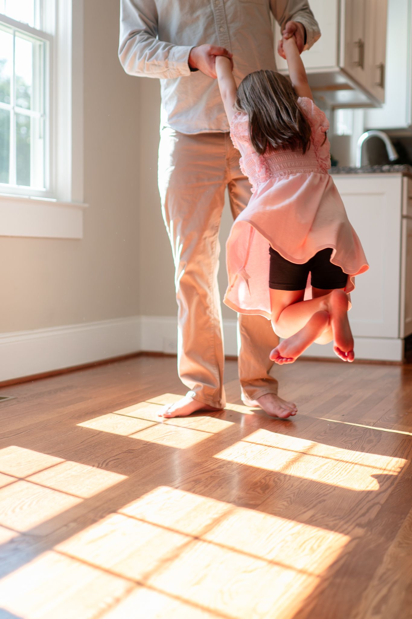 A small child in a pink sweater dances with an adult on a sunlit hardwood floor in a bright kitchen.