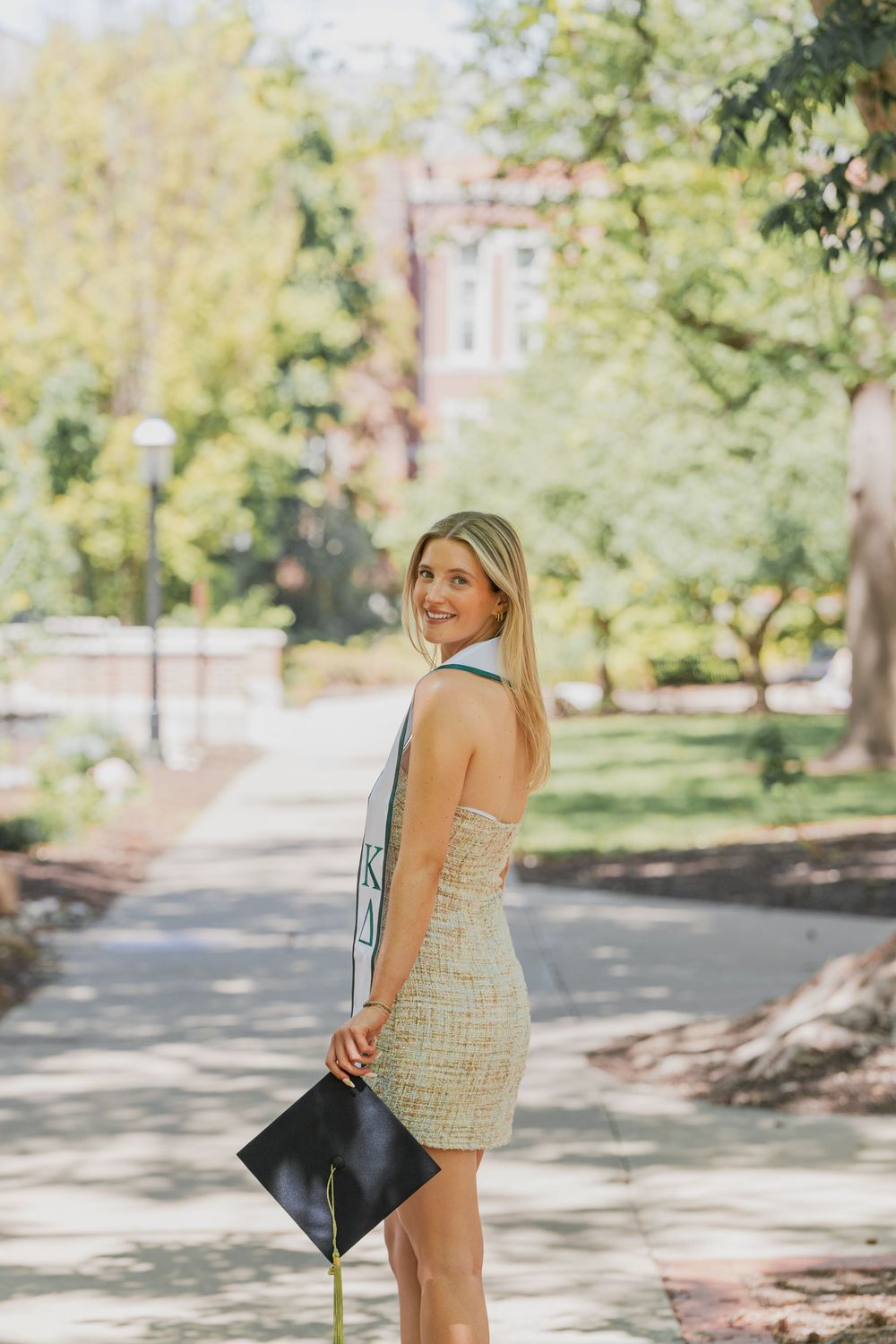 Smiling blonde Mizzou senior in tweed dress holding cap and sash on a sunny University of Missouri campus walkway.