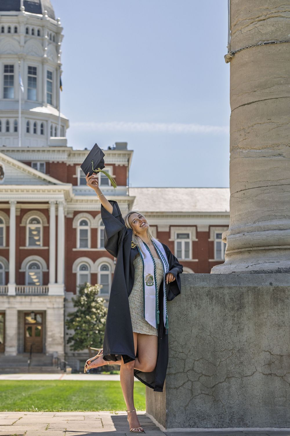 Mizzou graduate in cap and gown holding her cap aloft in front of a University of Missouri campus building.