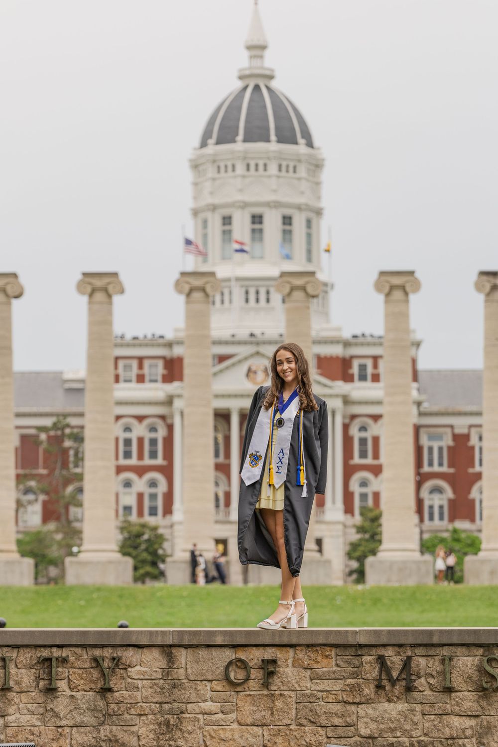 A smiling graduate in cap and gown stands on a stone wall in front of the University of Missouri columns and Jesse Hall.