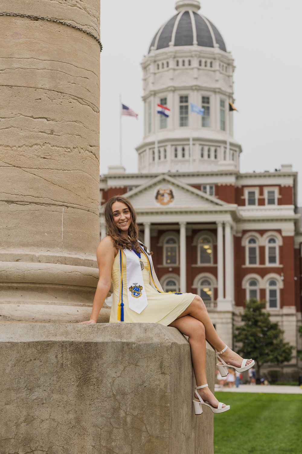 Mizzou graduate in yellow dress and white stole poses on a column base in front of Jesse Hall on the University of Missouri campus.