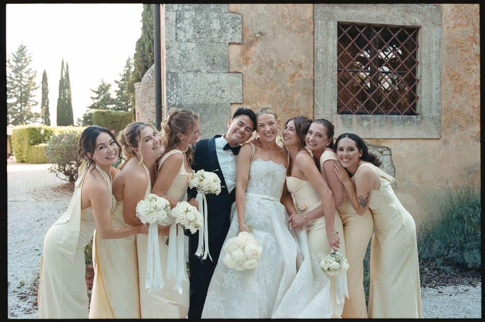 Bride and groom surrounded by smiling bridesmaids in cream dresses holding white bouquets outside a rustic stone building.