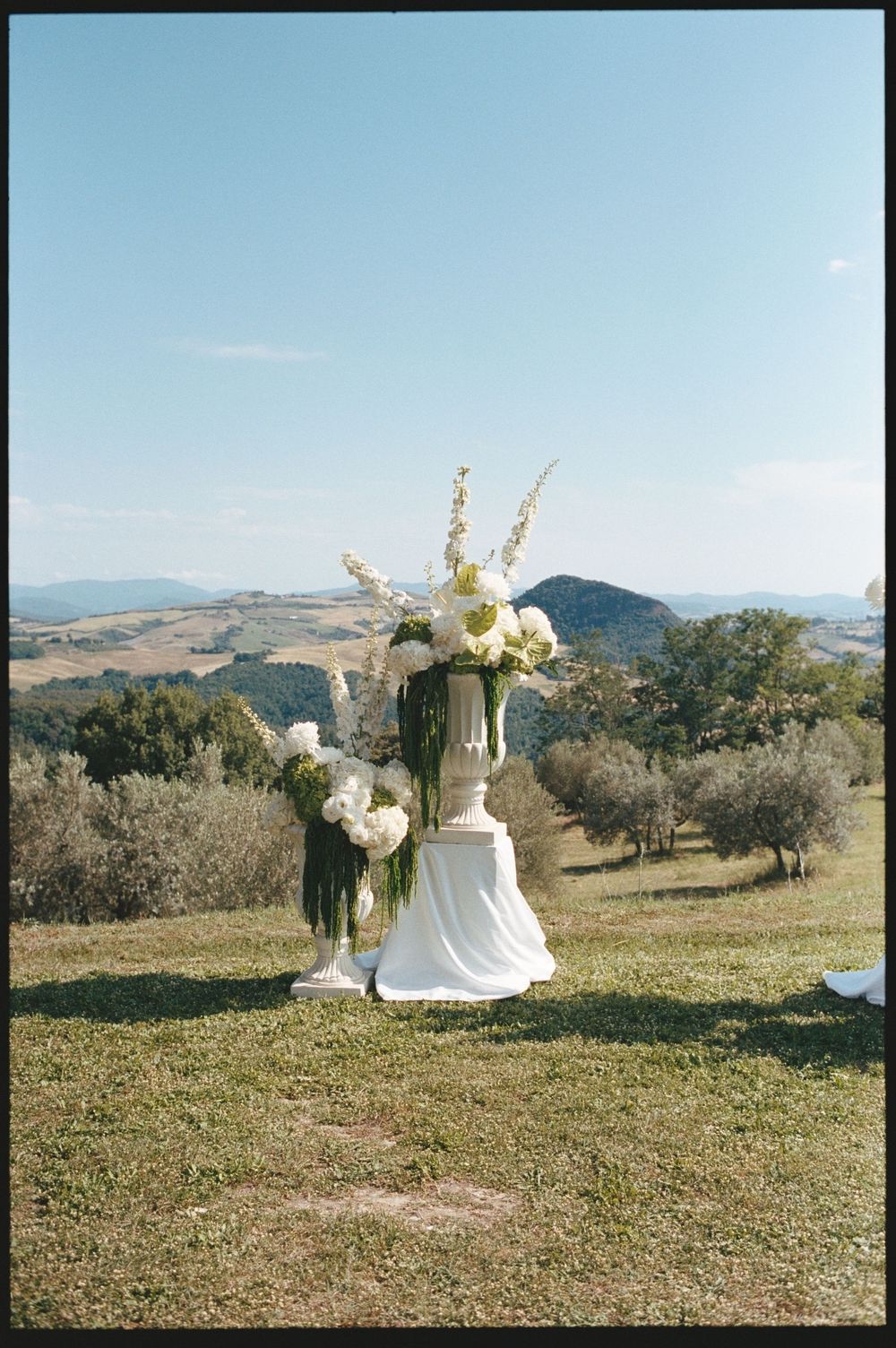 Elegant white floral arrangement in urn vase on draped pedestal with Tuscan hills backdrop at outdoor wedding ceremony.