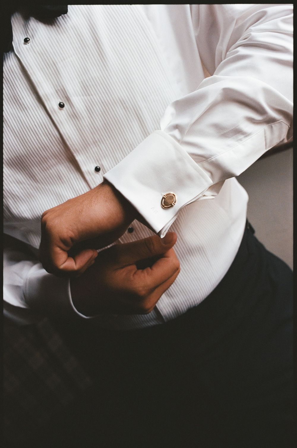 Man in white tuxedo shirt fastening gold cufflinks, close-up detail shot for formal wedding attire.