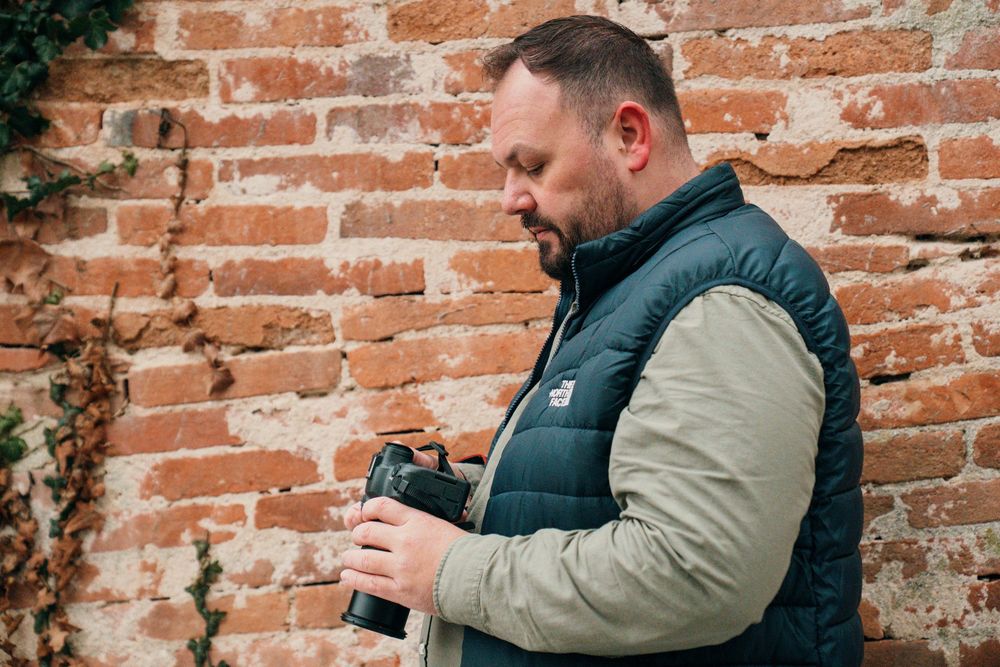 Person in a black vest and beige jacket uses smartphone while standing against rustic brick wall background.