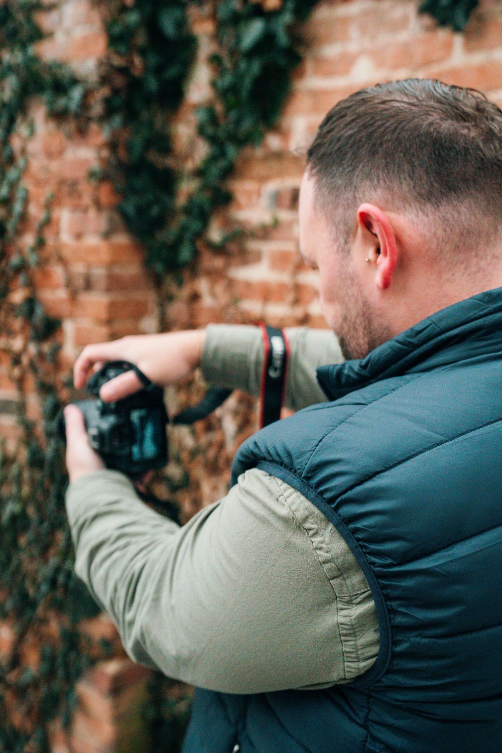 A photographer in a dark jacket and light sleeve aims a camera against a rustic brick wall backdrop.