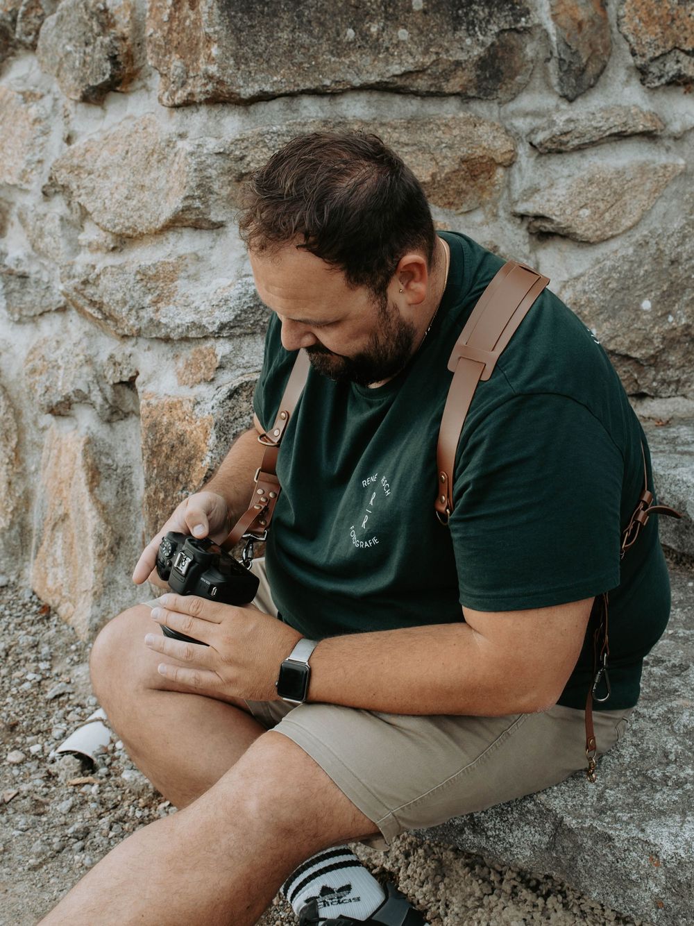 Man in green shirt sits against stone wall reviewing photos on his DSLR camera with leather strap.