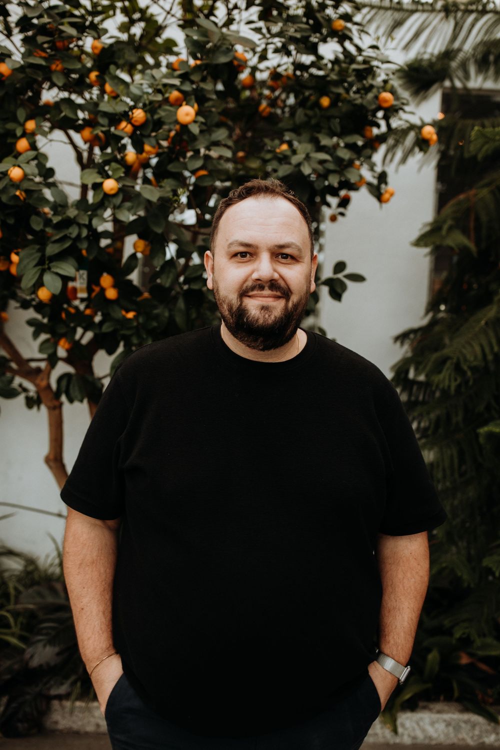 A person in a black t-shirt stands smiling in front of a citrus tree with orange fruits against a white wall.