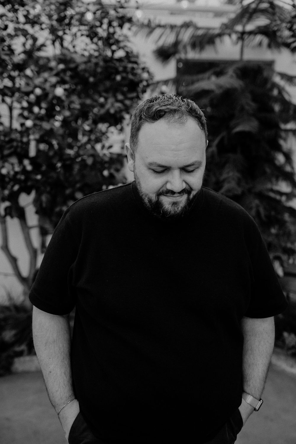 Black and white portrait of a person in a dark t-shirt looking down with a slight smile against a blurred outdoor background.