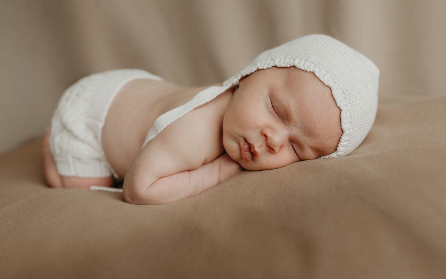 Peaceful sleeping newborn baby wrapped in white blanket on beige background in artistic newborn photography session.