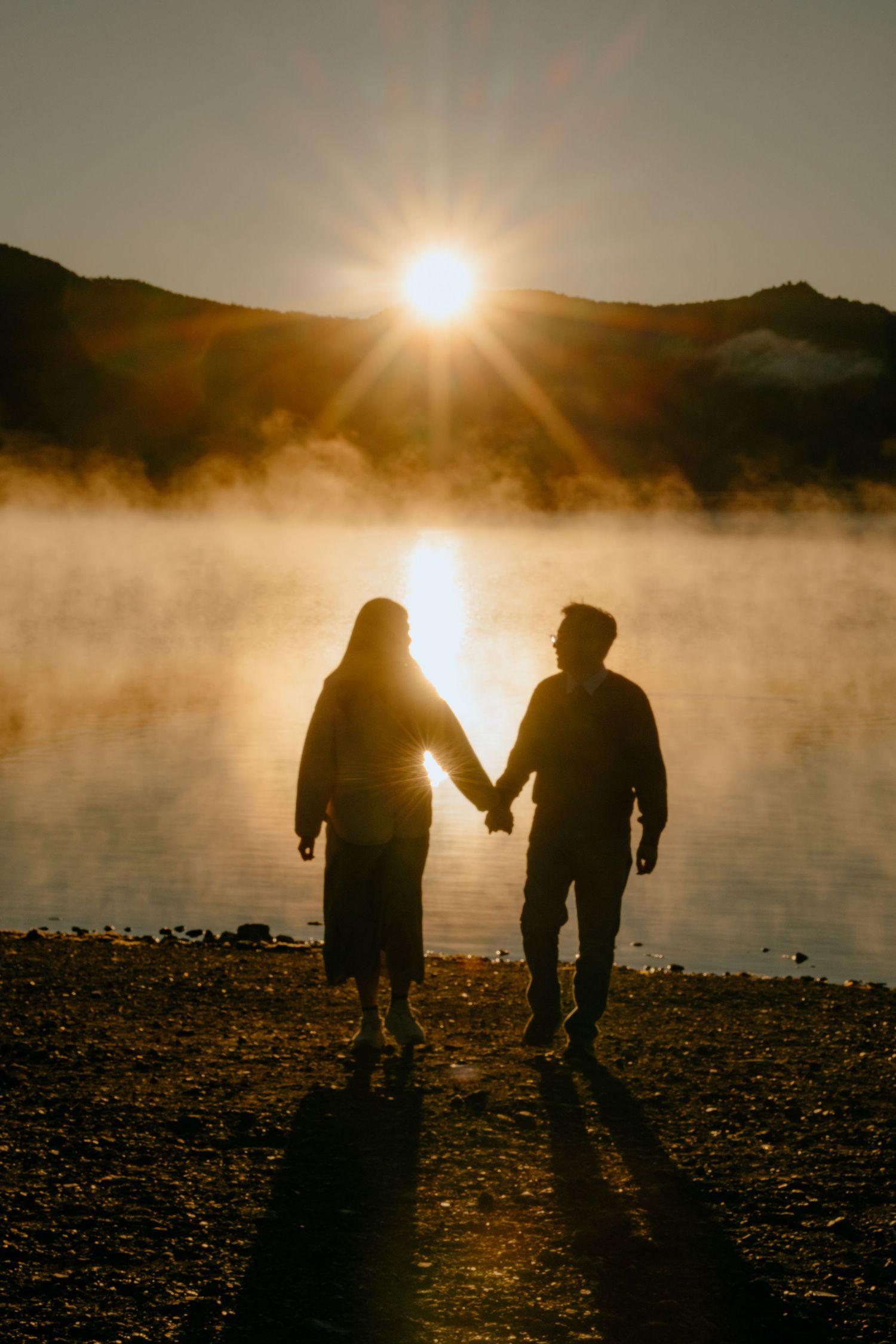 Silhouetted figures hold hands at sunset by a misty lake with sunburst effect creating a romantic atmosphere.