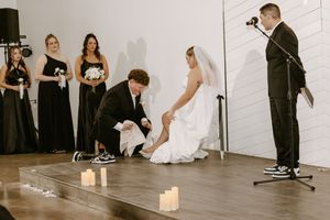 Groom kneeling to place shoes on bride during wedding ceremony with bridesmaids in black dresses standing nearby.