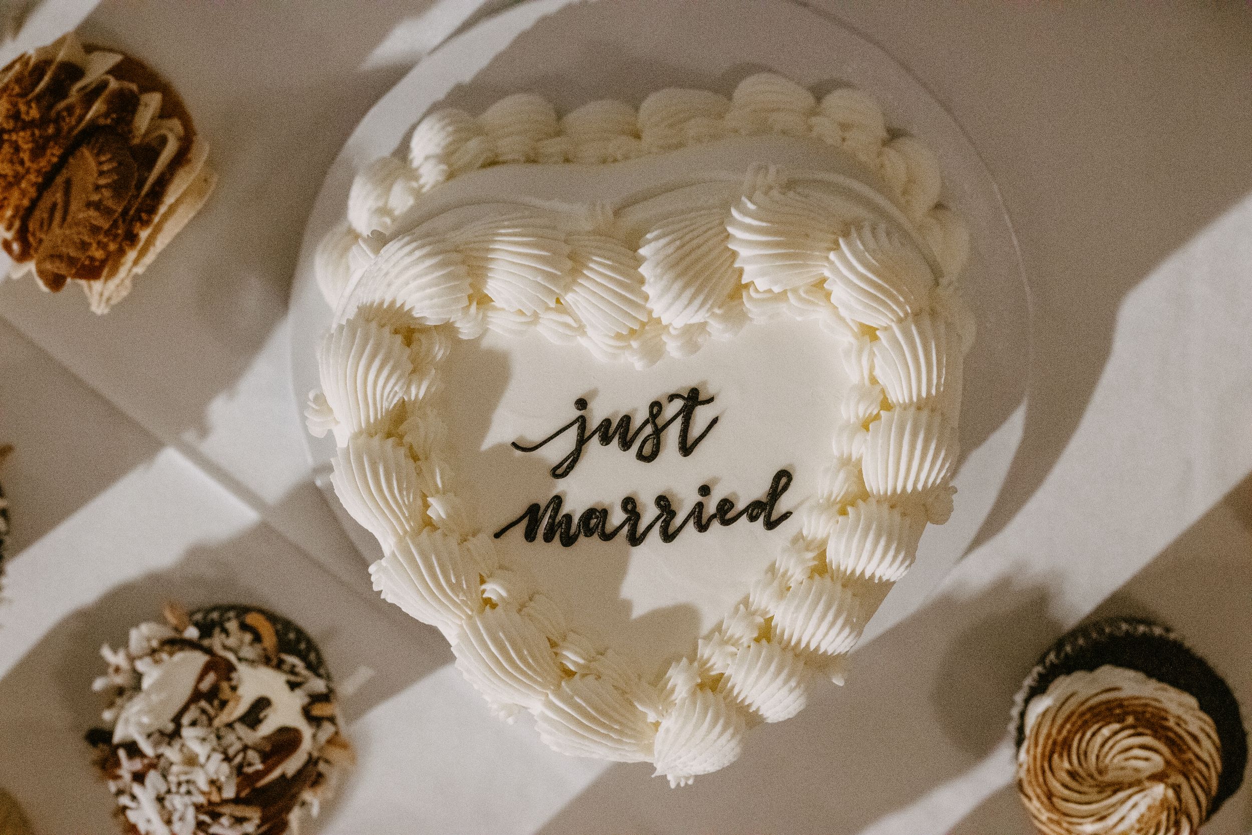Heart-shaped white wedding cake decorated with "just married" script, surrounded by cupcakes on a white table.