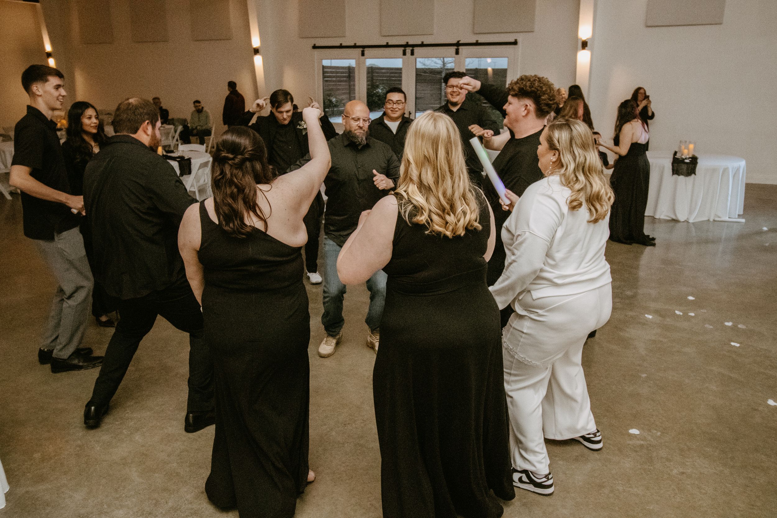 Guests dancing together at a wedding reception in a modern venue, dressed in elegant black formal attire.