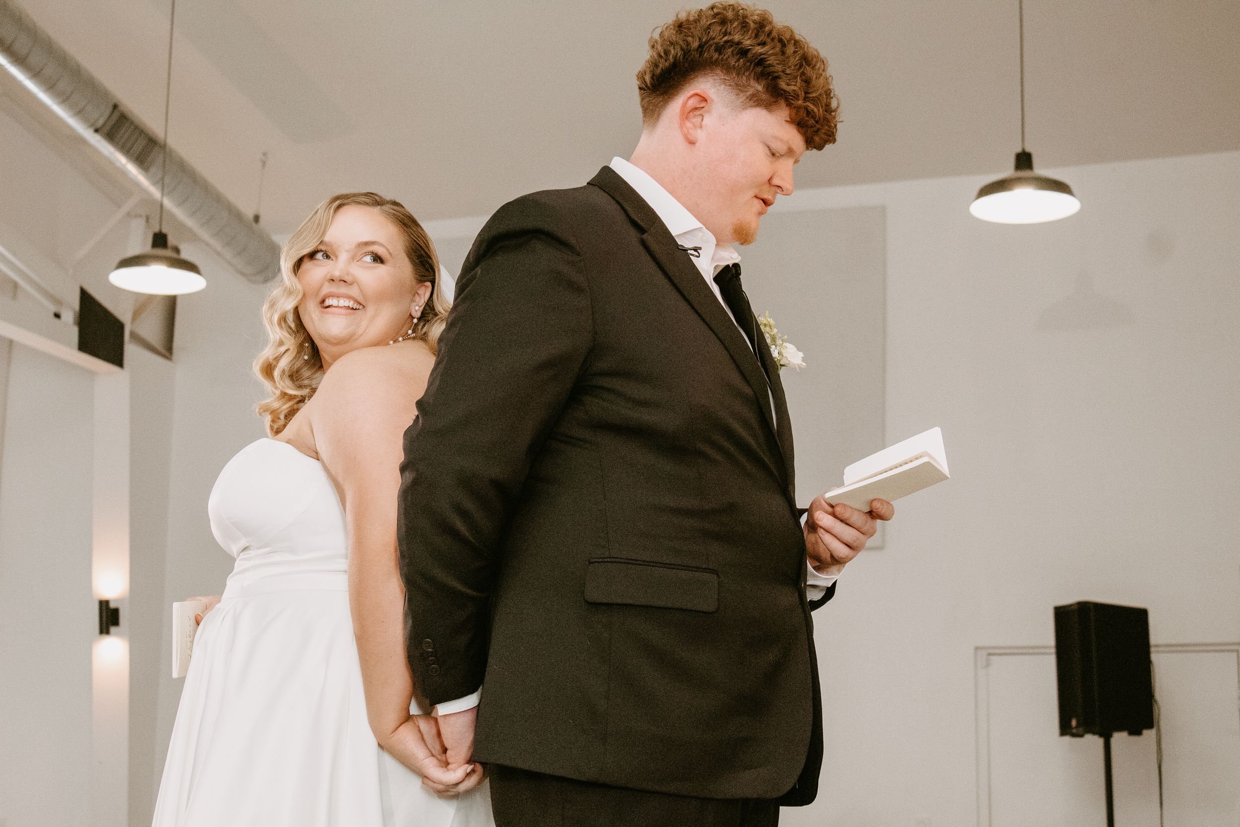 Bride and groom stand back-to-back holding hands, reading vows at indoor wedding ceremony.
