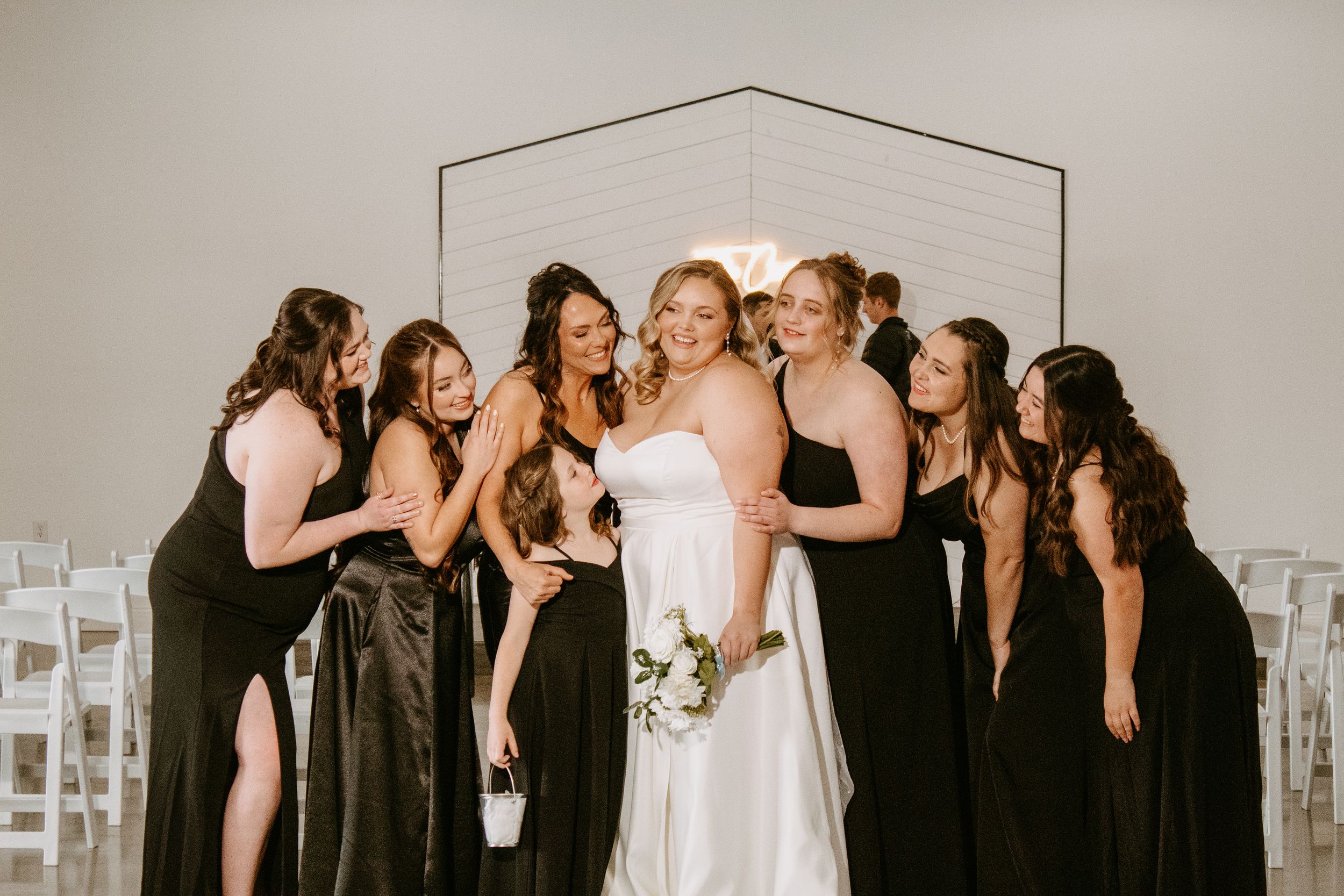Bride in white gown surrounded by bridesmaids in black dresses, laughing together before a geometric arch backdrop.
