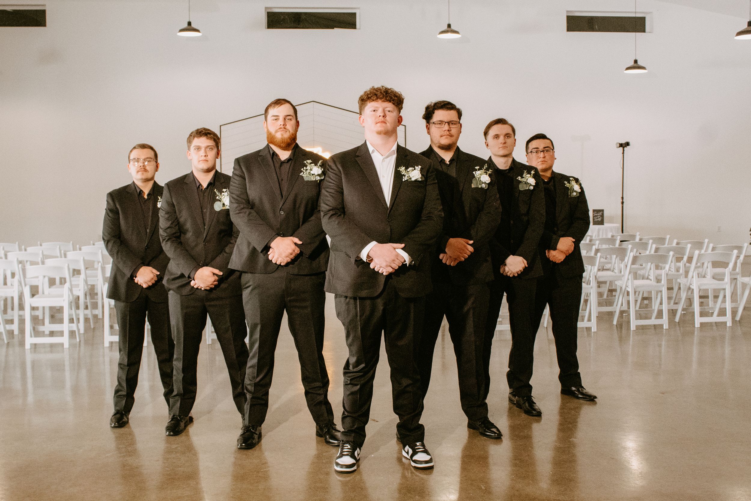 Groom stands with six groomsmen in black suits with floral boutonnieres in a white wedding venue.