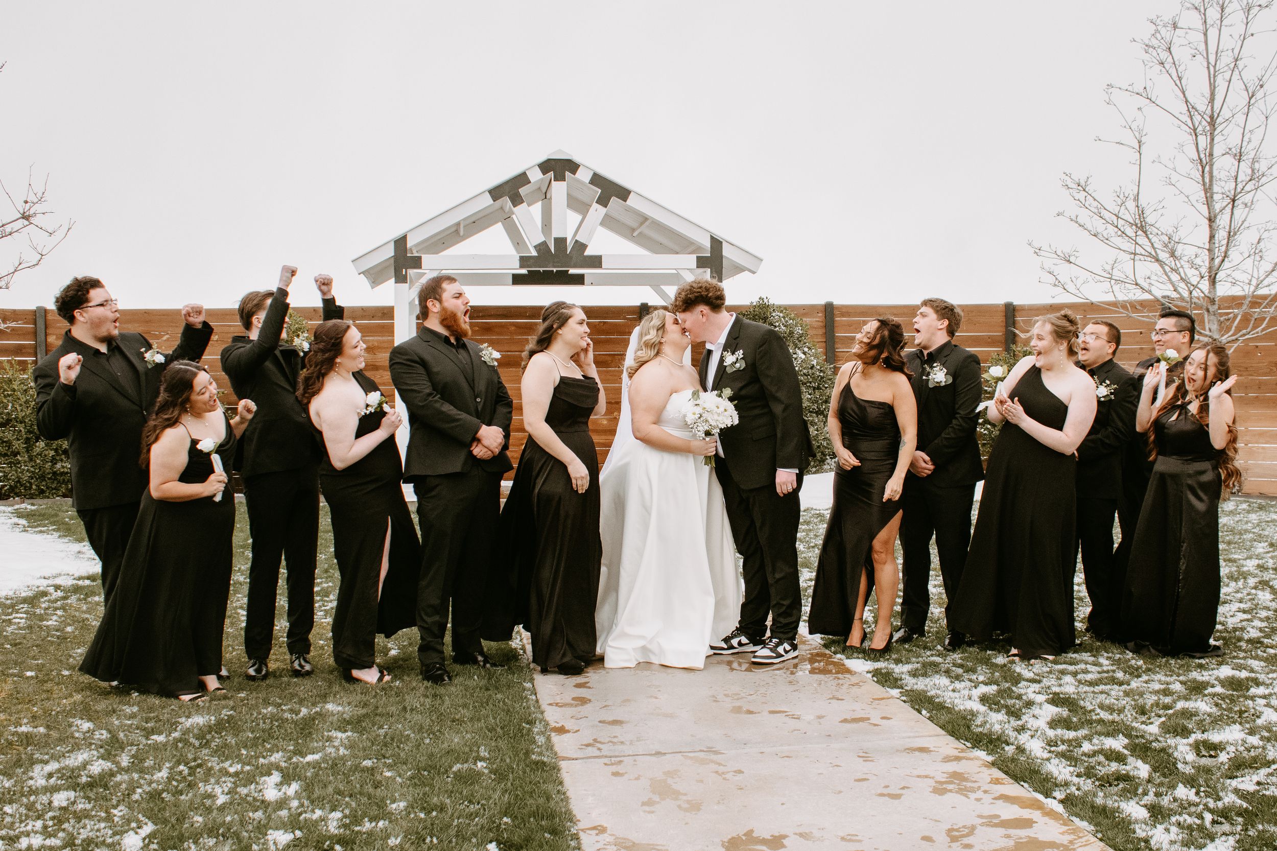 Wedding party in black attire celebrates with bride and groom kissing outdoors in snowy winter setting.