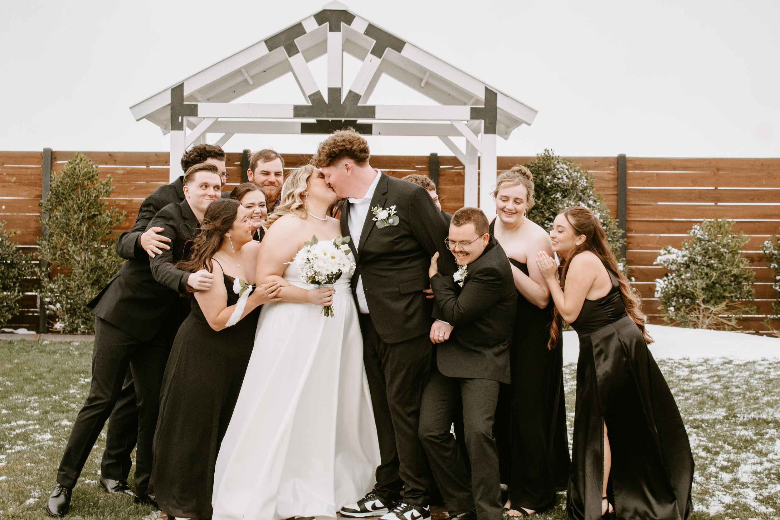 Wedding party celebrates with bride and groom kissing under white arbor, guests in black attire cheering outdoors.