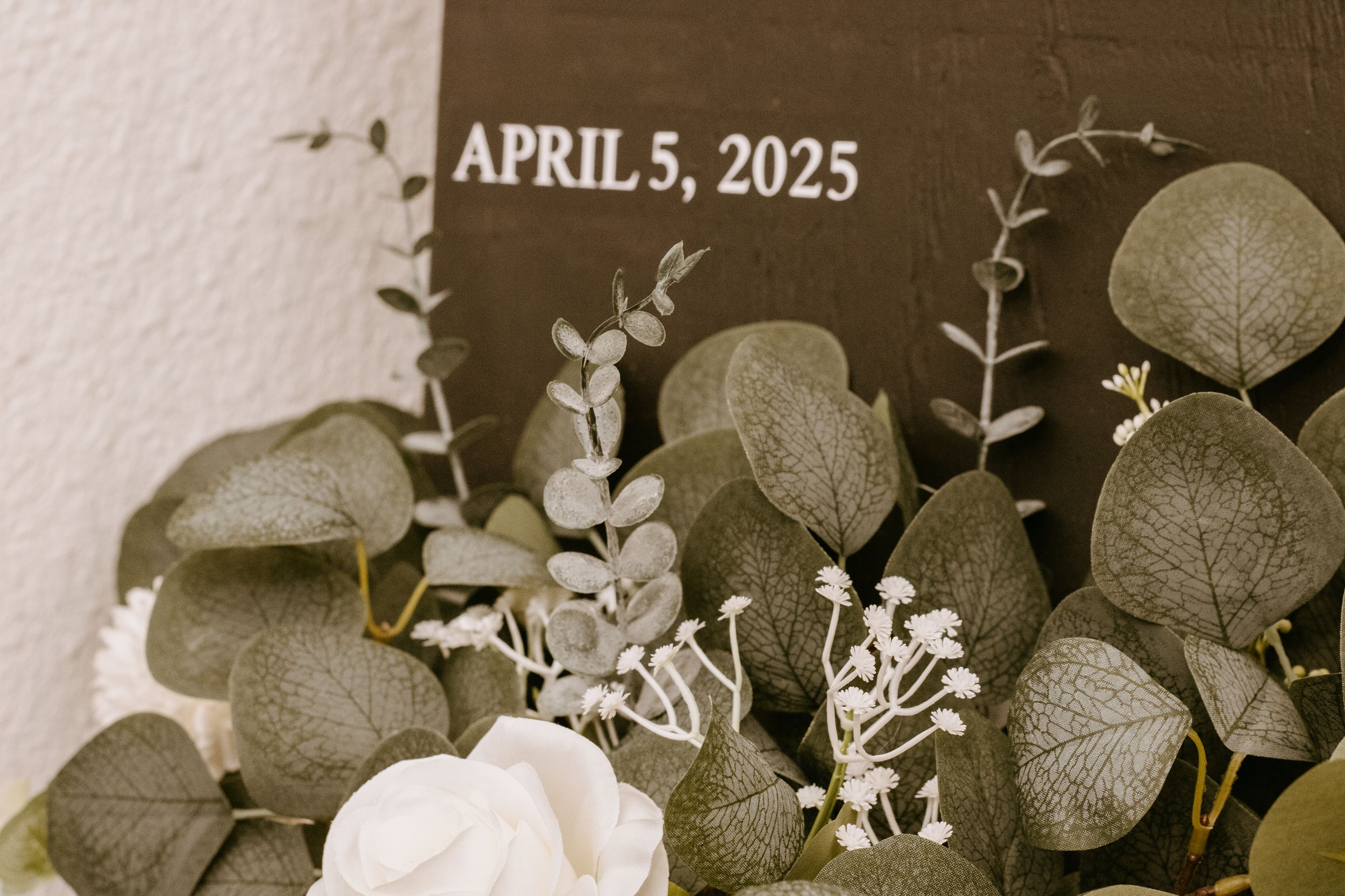 Dark sign reading April 5, 2025 surrounded by elegant eucalyptus leaves and white roses for a wedding decor display.