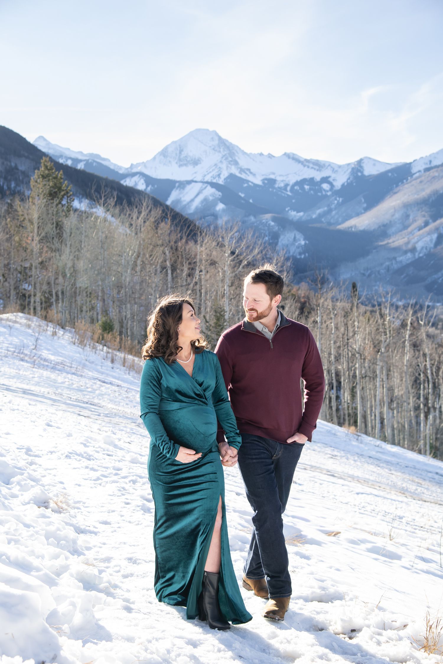 A couple walks together in formal attire through snowy mountains with aspen trees and peaks in the background for maternity photos Colorado