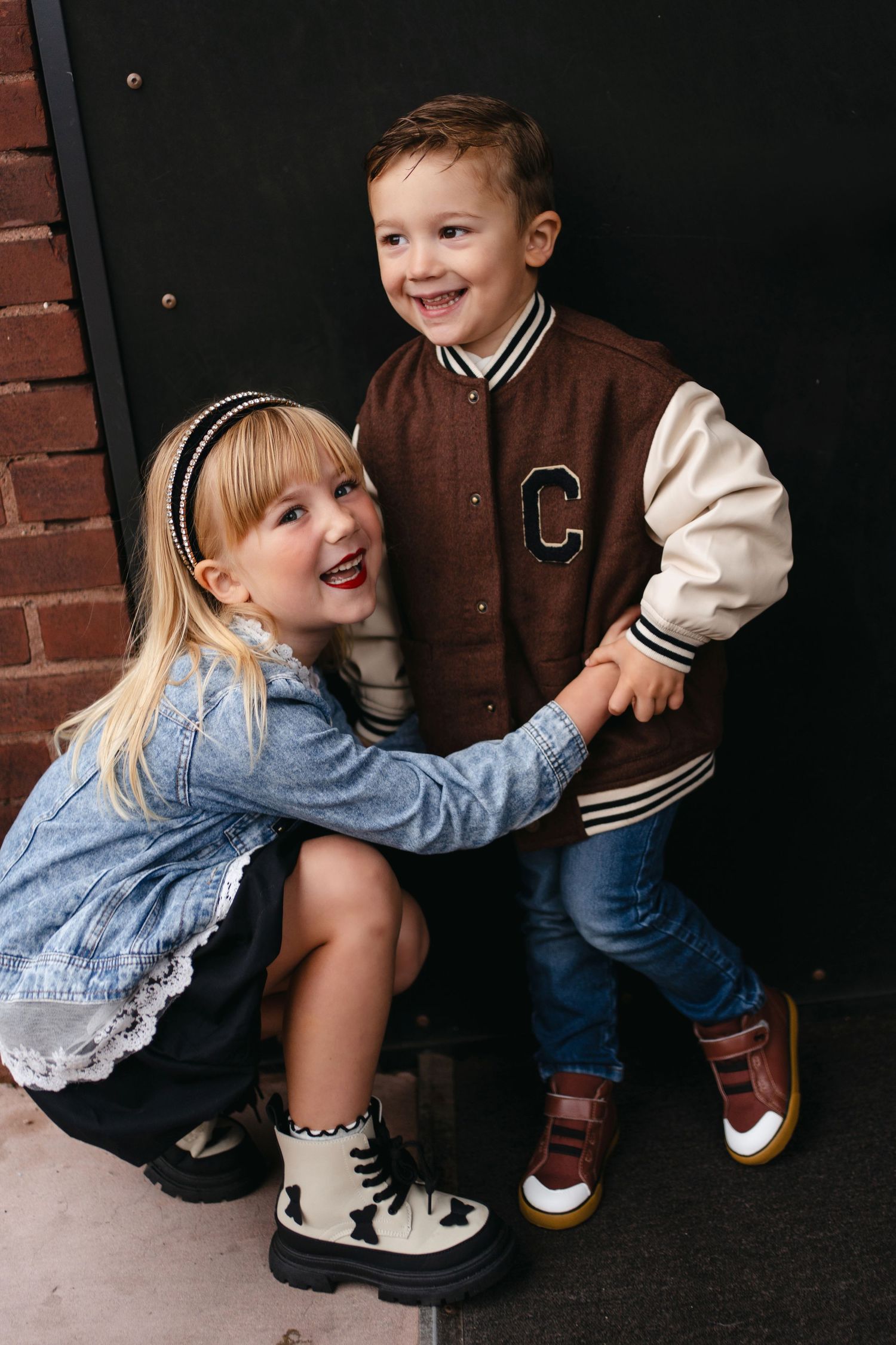 Two children in casual clothing and sneakers crouch playfully against a dark door and brick wall background.