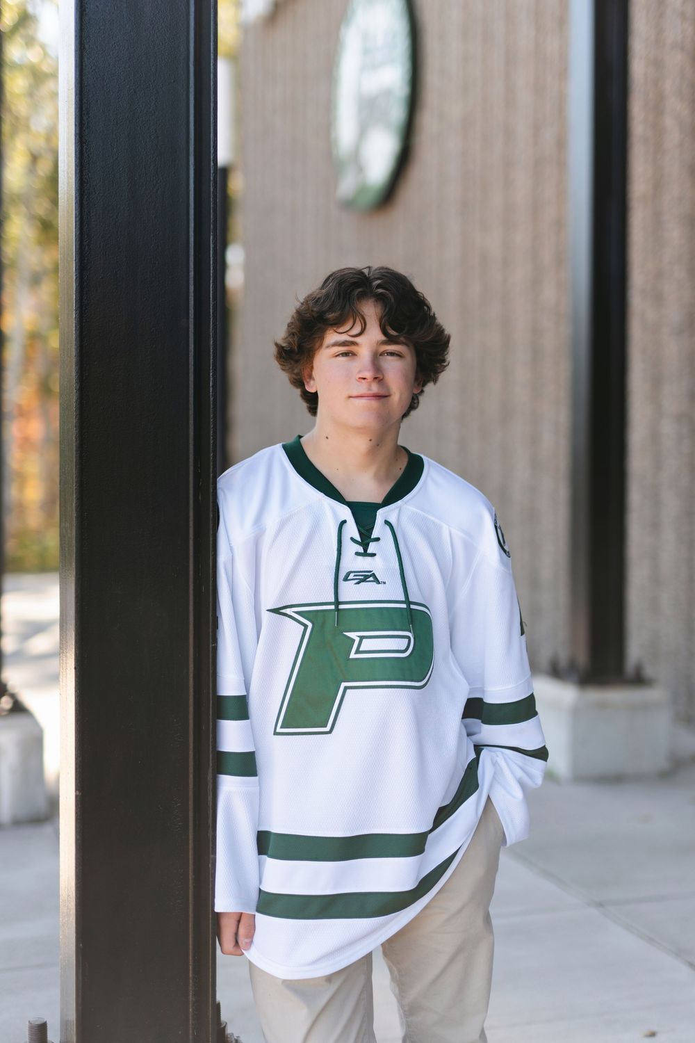 Student athlete poses in white and green P-logo hockey jersey against building exterior.