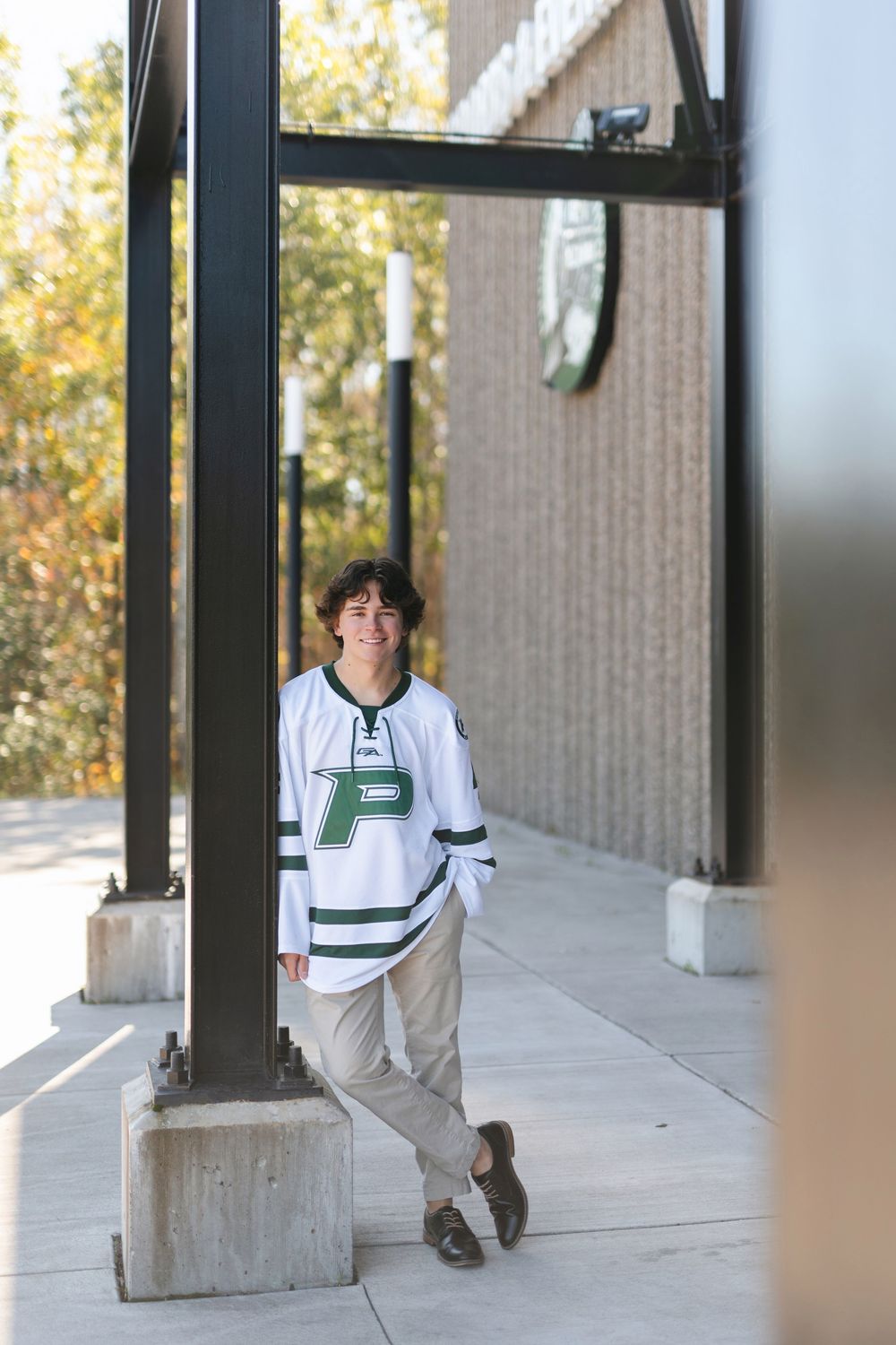Student in a white hockey jersey leans against a pillar outside a building on a sunny day.