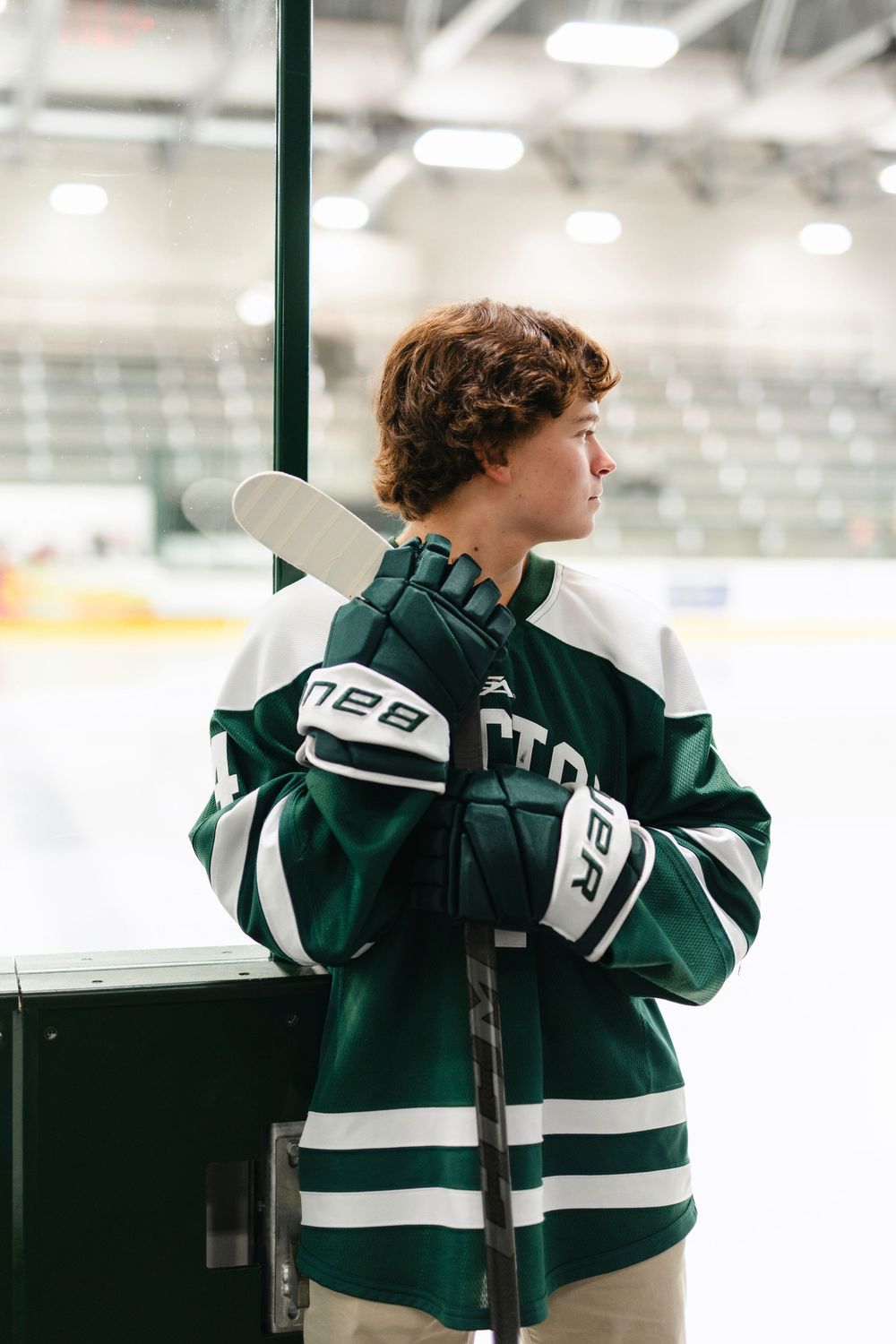 Hockey player in green and white jersey leans against rink boards while holding stick in indoor arena.