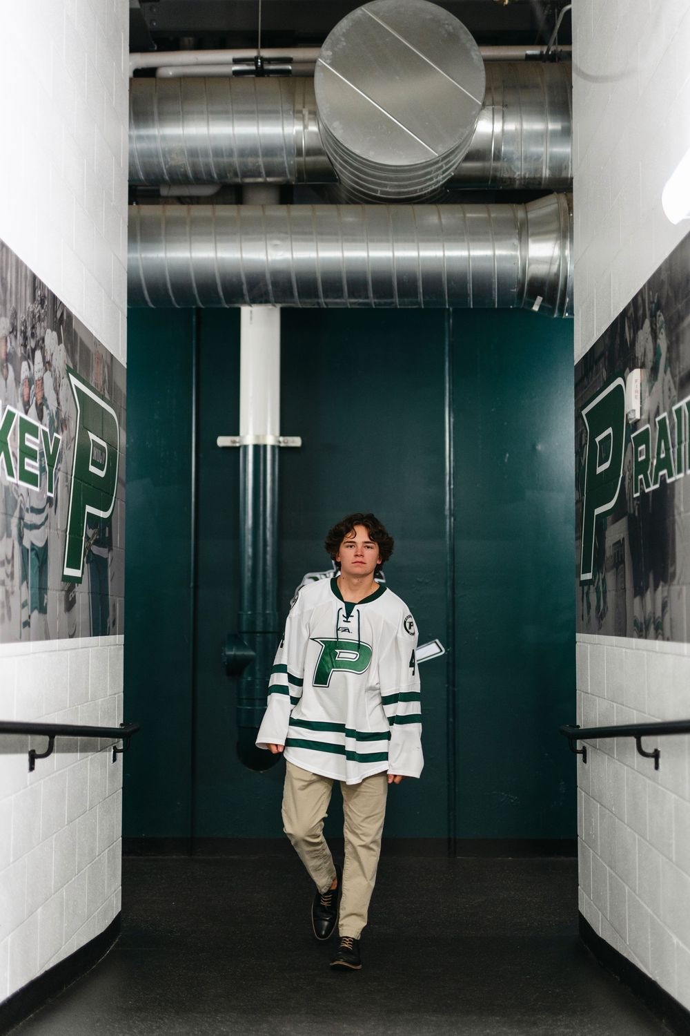 Person wearing a white and green sports jersey walks through a stadium tunnel with ductwork overhead.