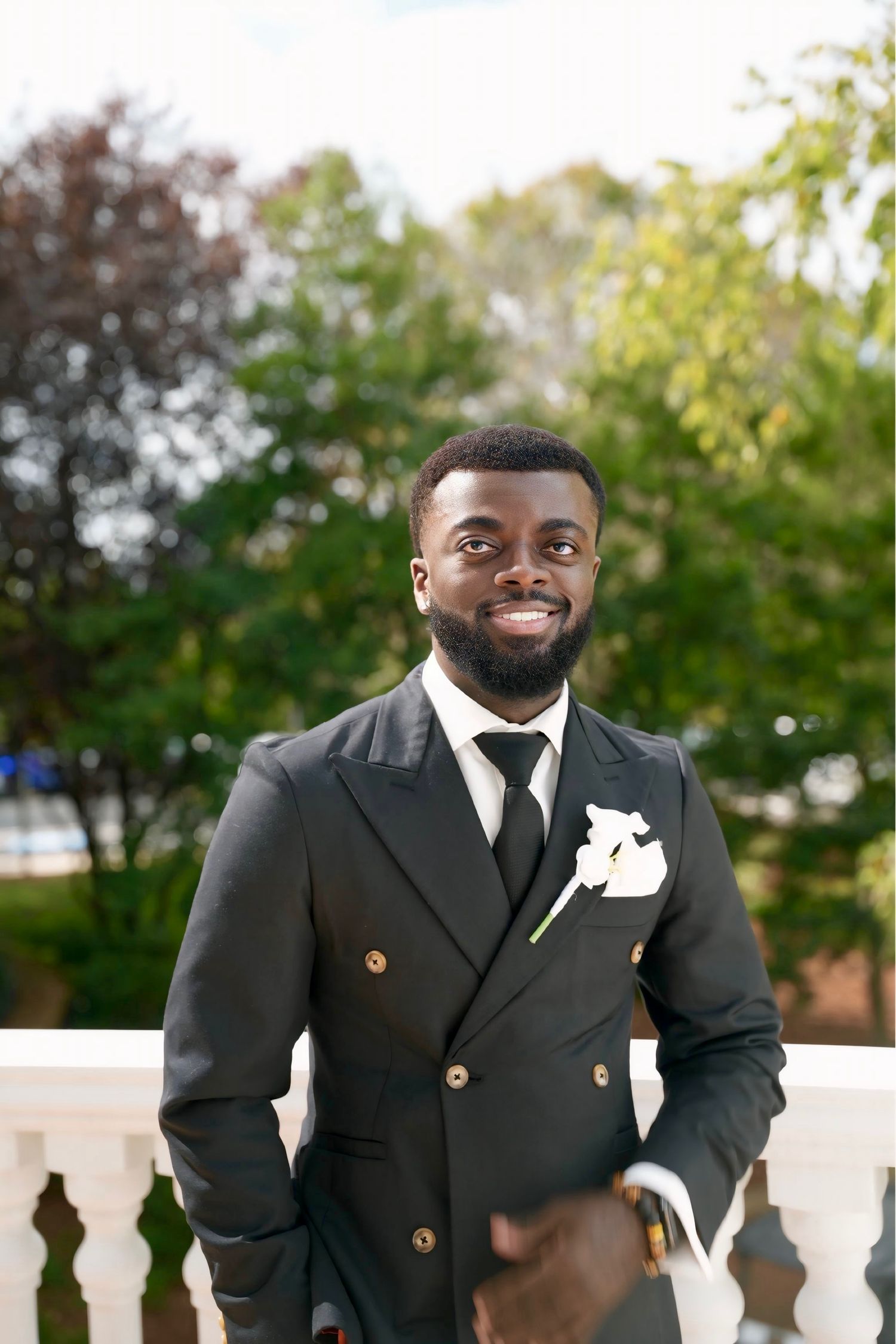 A groom in a black suit with a white boutonniere smiles warmly during an outdoor wedding celebration.