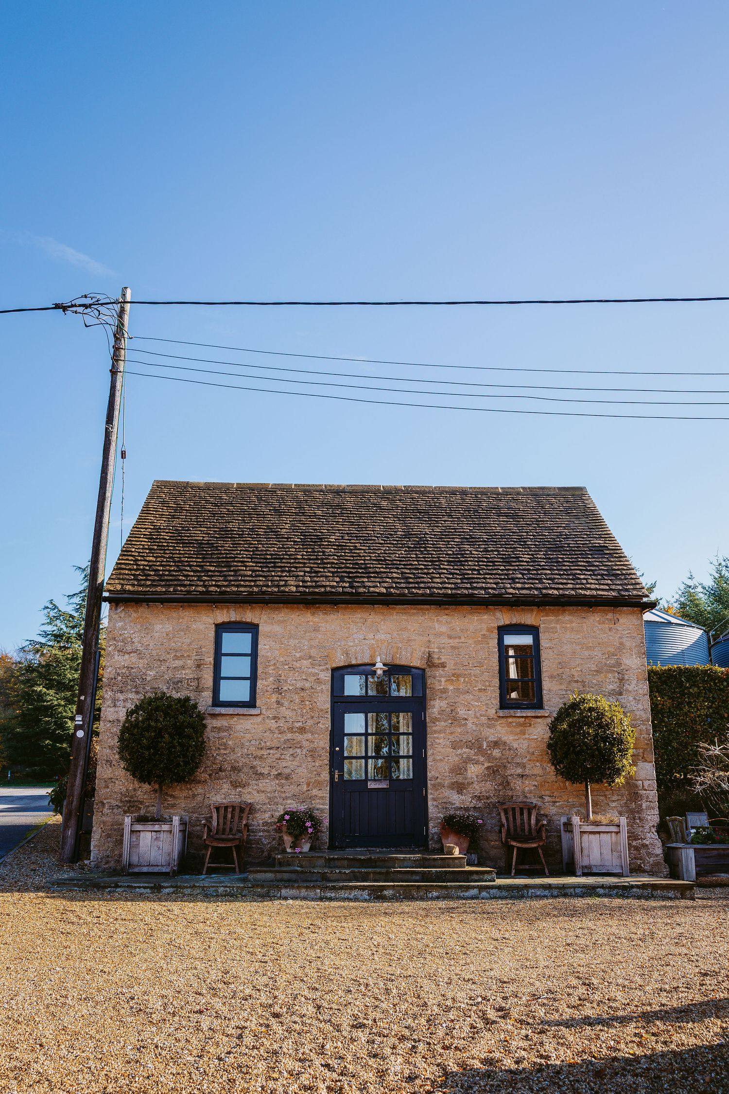 Charming rustic stone cottage with blue door and potted topiary plants stands against clear blue sky with power lines overhead.