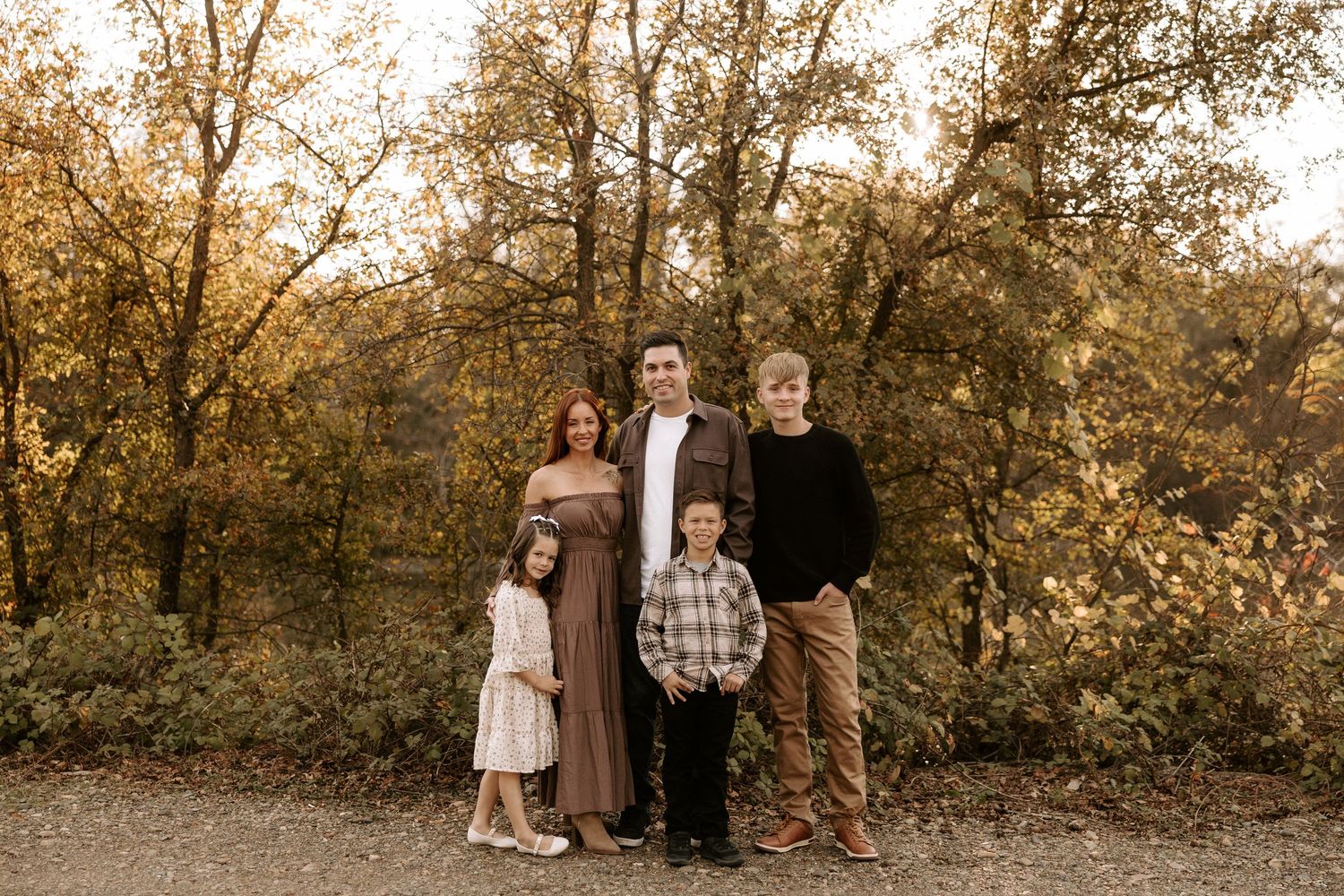 A family group stands together on a path surrounded by autumn trees with golden leaves in warm evening light.